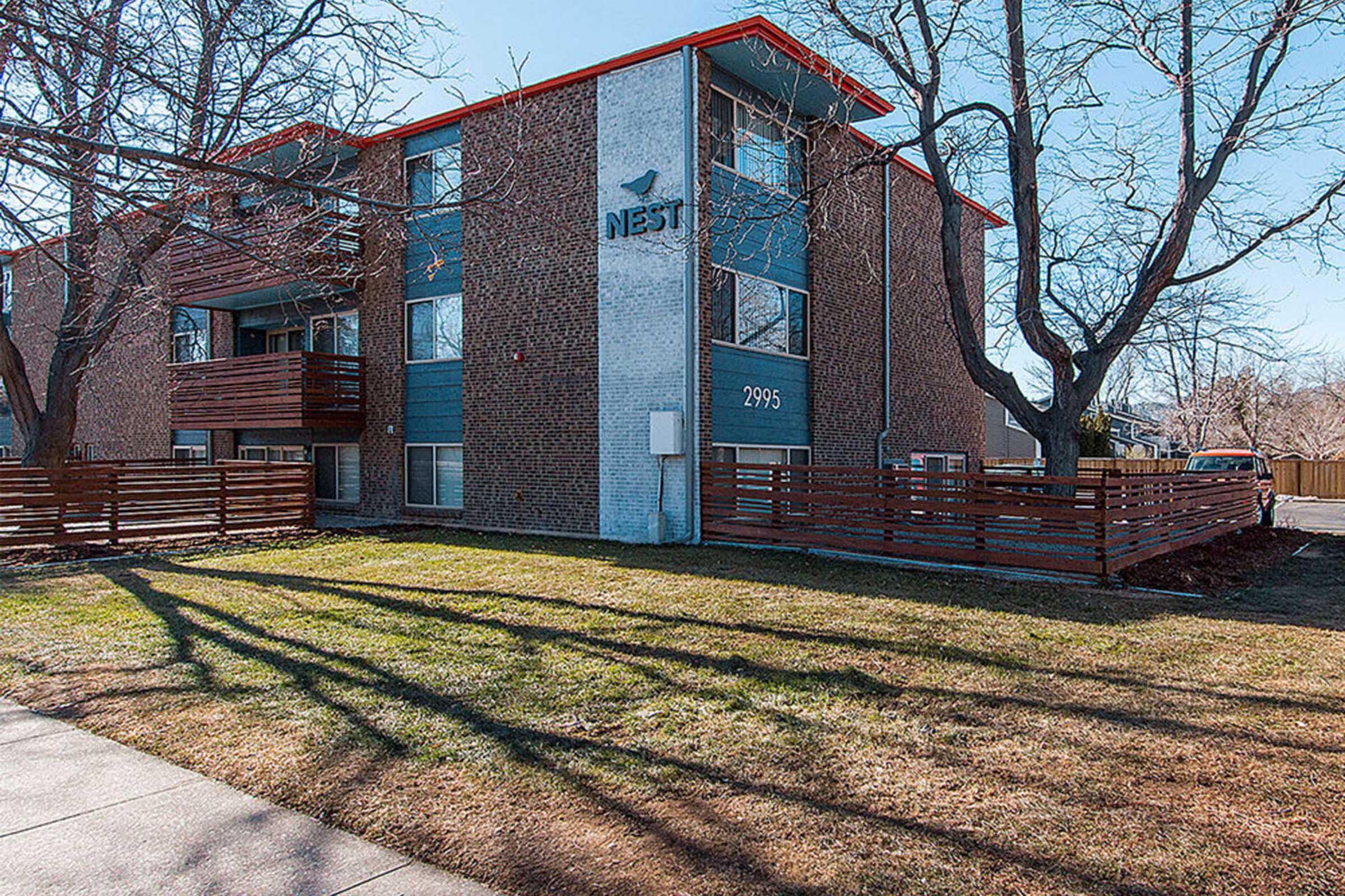 A brick apartment building with the sign "NEST" displayed prominently on the front. The building has two stories, balconies with wooden railings, and a well-maintained lawn with shadows from nearby trees. The number "2995" is visible on the building's facade.