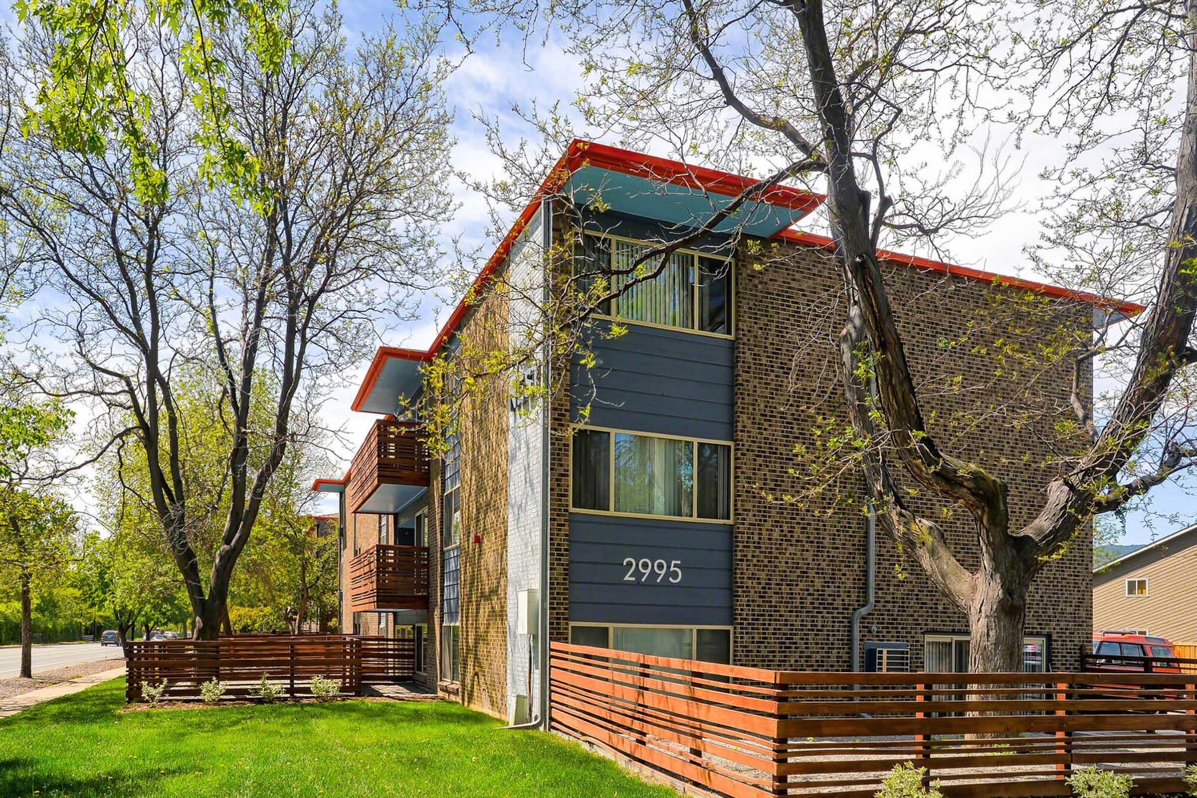 A modern brick apartment building with a gray facade and red roof accents, featuring balconies and large windows. Surrounding the building are green trees and a grassy area, with wooden fencing visible in the foreground. The building is located on a tranquil street.