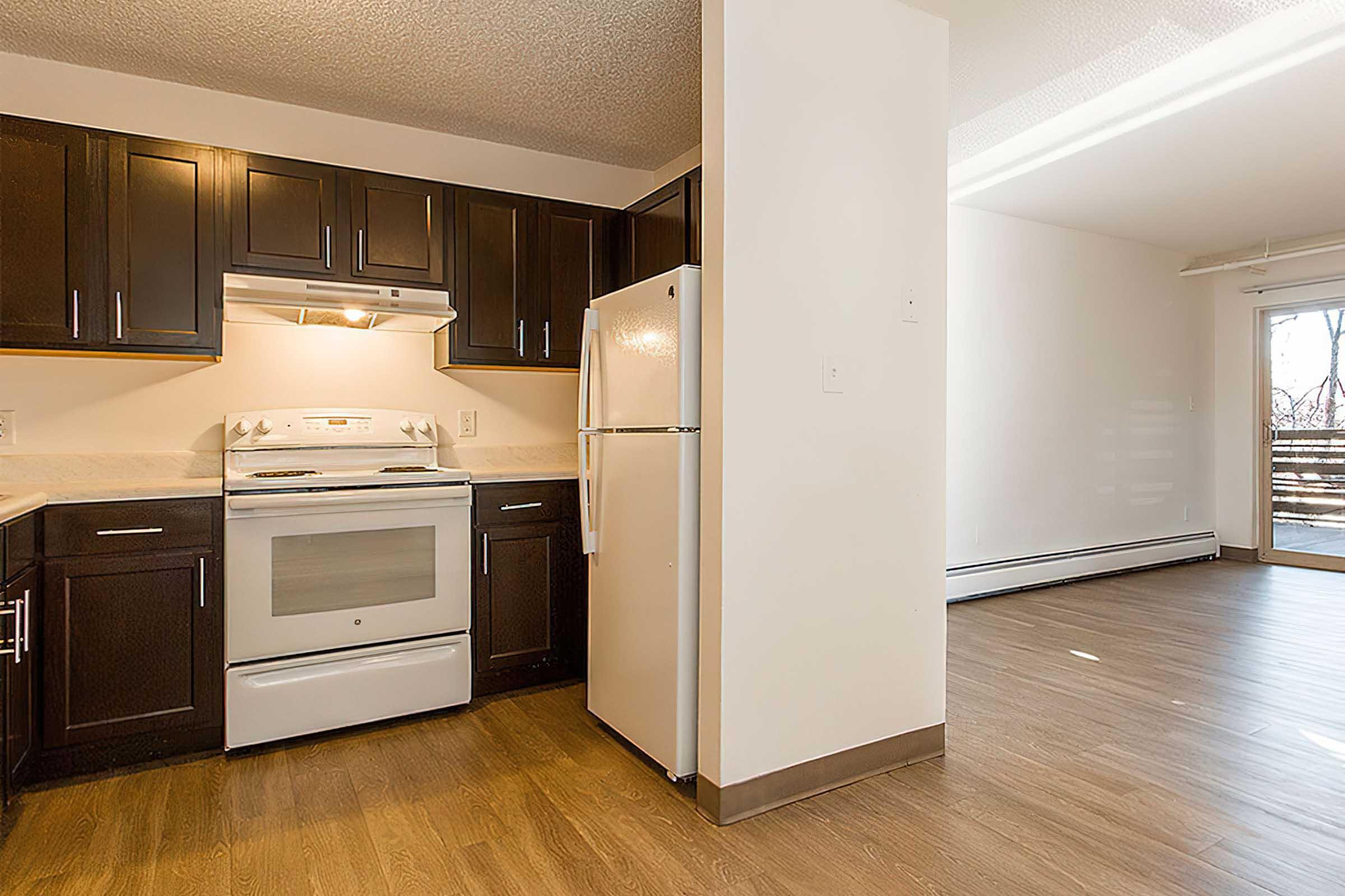 A modern kitchen featuring dark wooden cabinetry, a white stove and refrigerator, and light-colored countertops. The room has an open layout with a spacious living area visible in the background, complete with natural light streaming in through large windows.