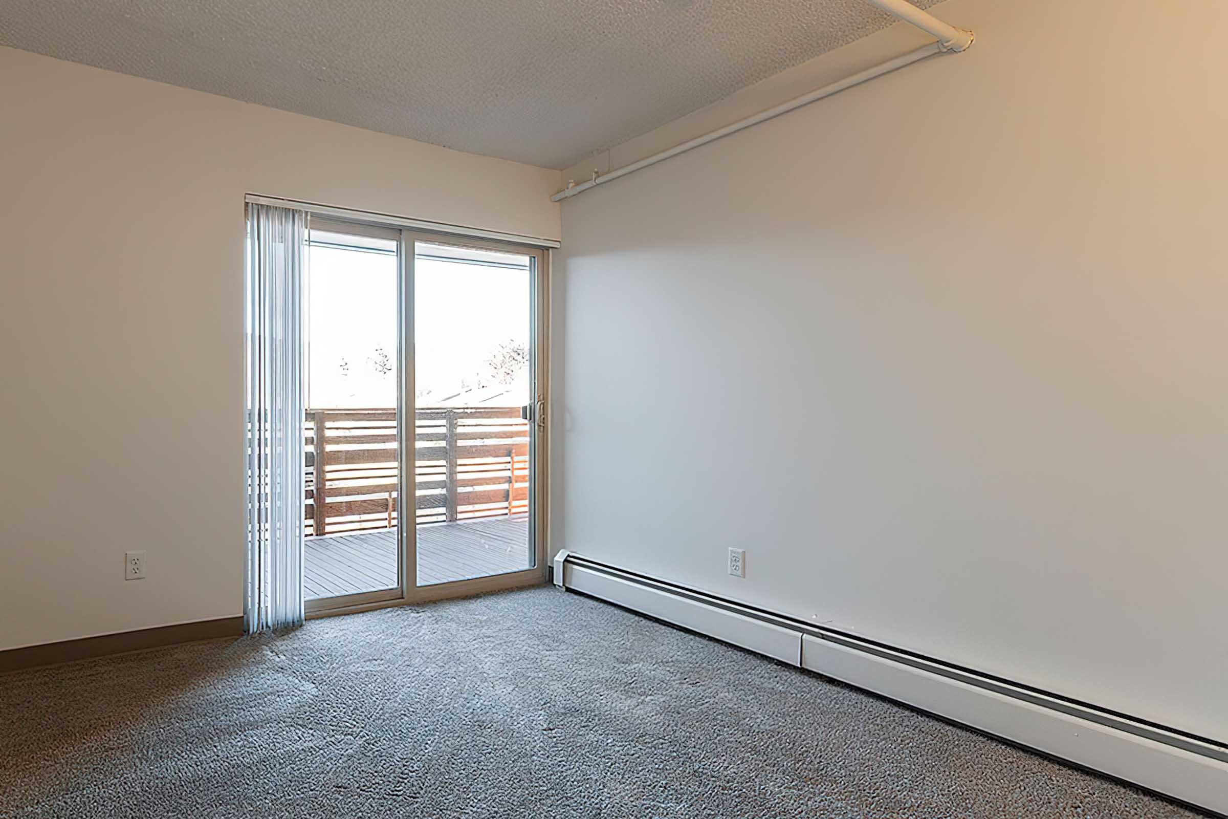 A bright, empty room with a light-colored wall and carpeted floor. A sliding glass door leads to a wooden balcony, providing natural light. The room has minimal furnishings, creating a spacious and open feel.