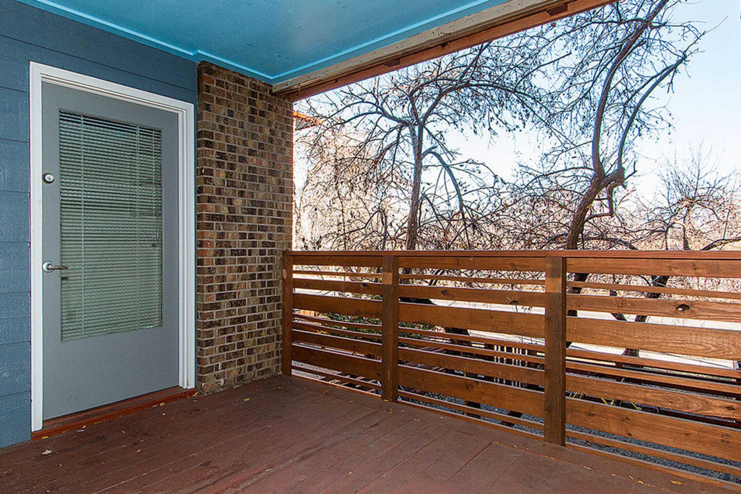 A partially shaded balcony featuring a wooden railing and an open door. The floor is wooden, and outside, trees can be seen through the railing. The walls are a mix of blue and brick, contributing to a cozy outdoor space.