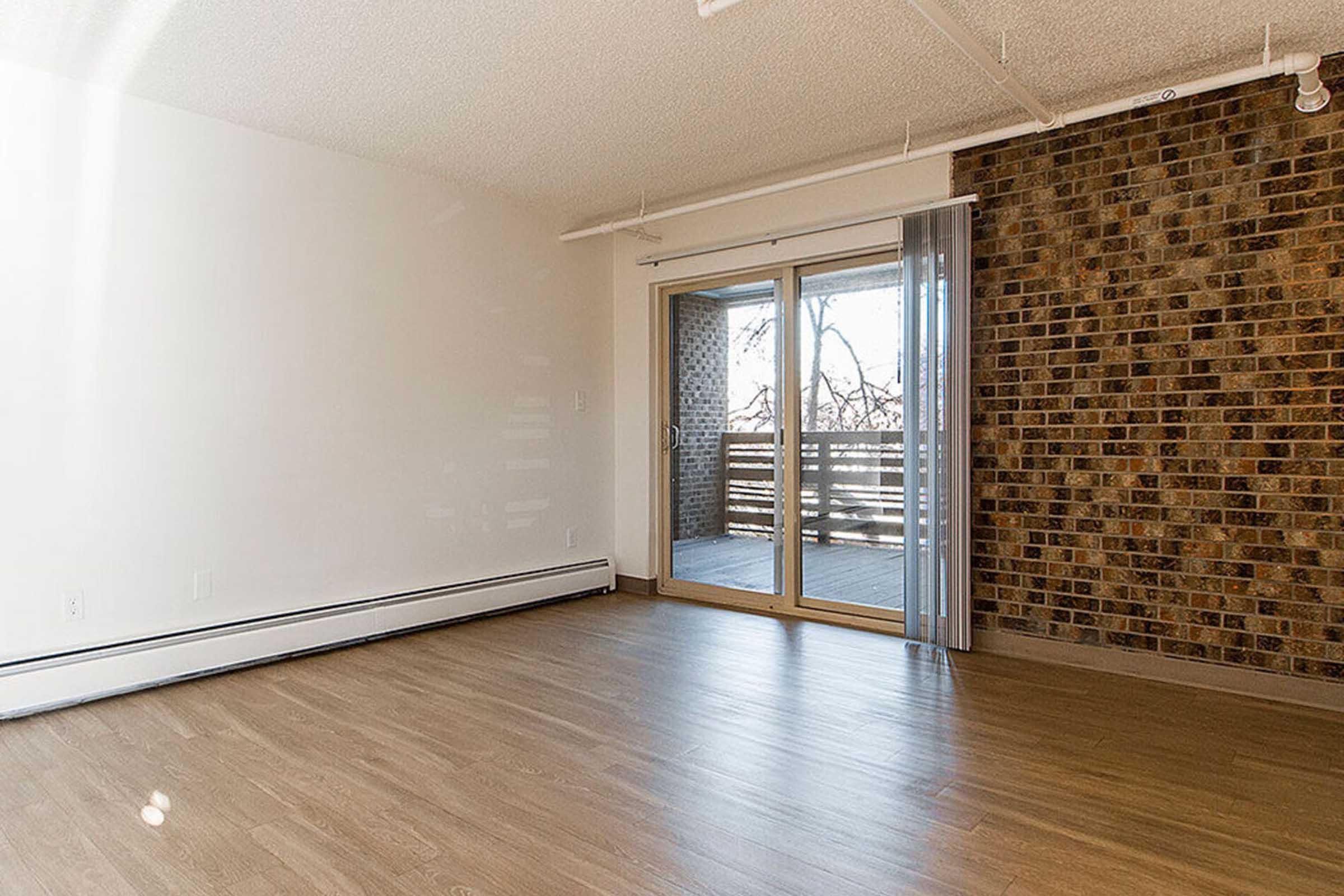 A spacious, empty room with a light-colored, textured wall and a brick feature wall. Large sliding glass doors open to a balcony area, allowing natural light to fill the space. The flooring is a warm, wood-like design, contributing to a modern and inviting atmosphere.