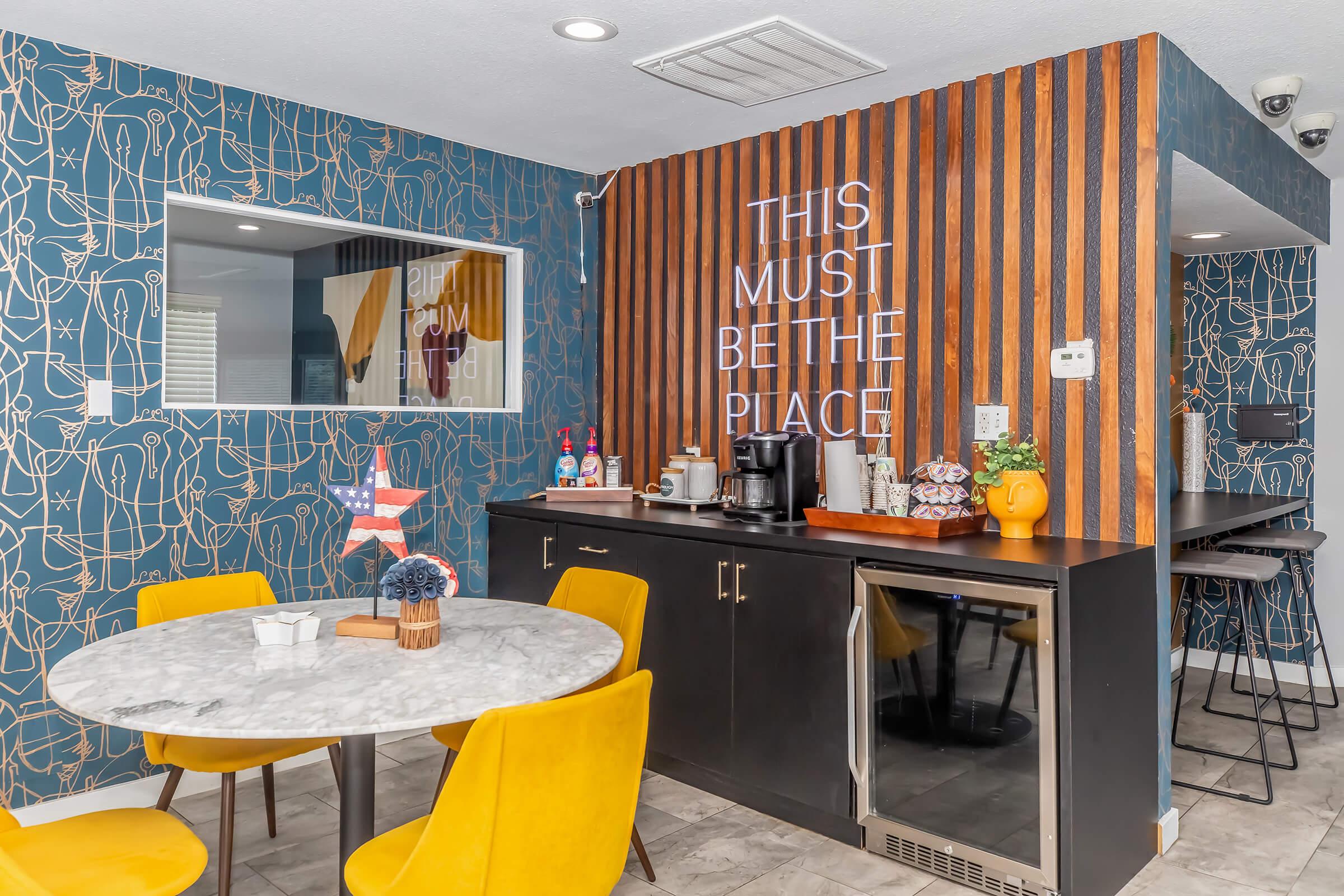 A modern kitchen area featuring a marble dining table with yellow chairs. There is a dark countertop with a coffee maker and a beverage fridge. The wall behind has wooden paneling with the phrase "THIS MUST BE THE PLACE" illuminated, while the other wall is adorned with a blue patterned wallpaper.