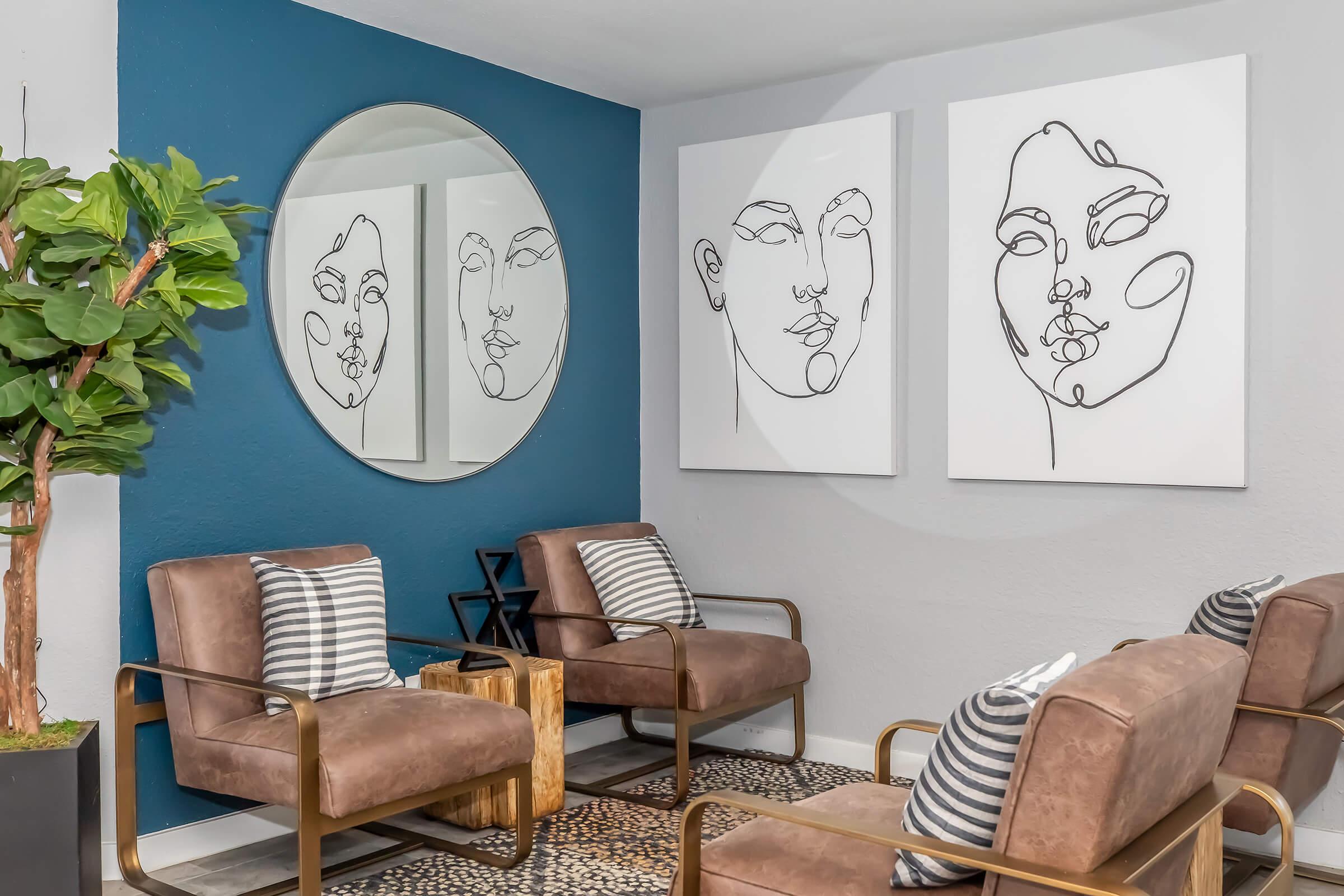 A modern seating area featuring two abstract line drawings of faces on white canvases. There's a large round mirror on a blue accent wall, two stylish brown chairs with gold frames, and greenery in a decorative pot. The floor is covered with a patterned rug, creating an inviting and contemporary atmosphere.