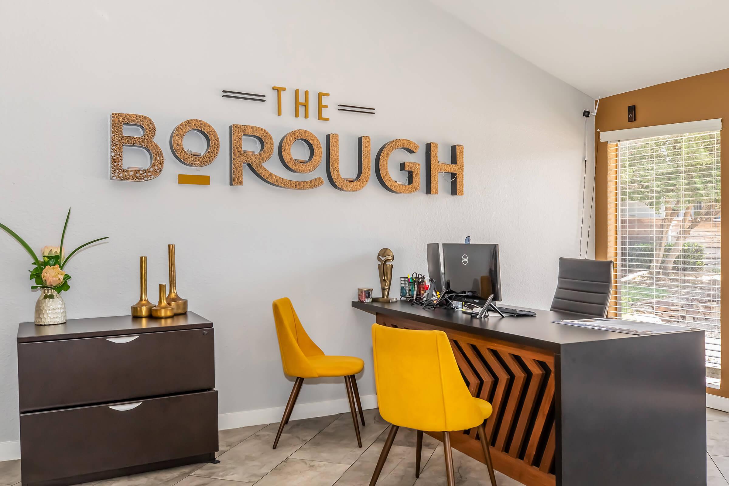 A modern office reception area featuring a large sign that reads "THE BOROUGH" on a light gray wall. There is a stylish wooden desk with a computer, black office chair, and two bright yellow chairs. Decor includes a flower arrangement and decorative items on the desk and a drawer unit. Large windows let in natural light.