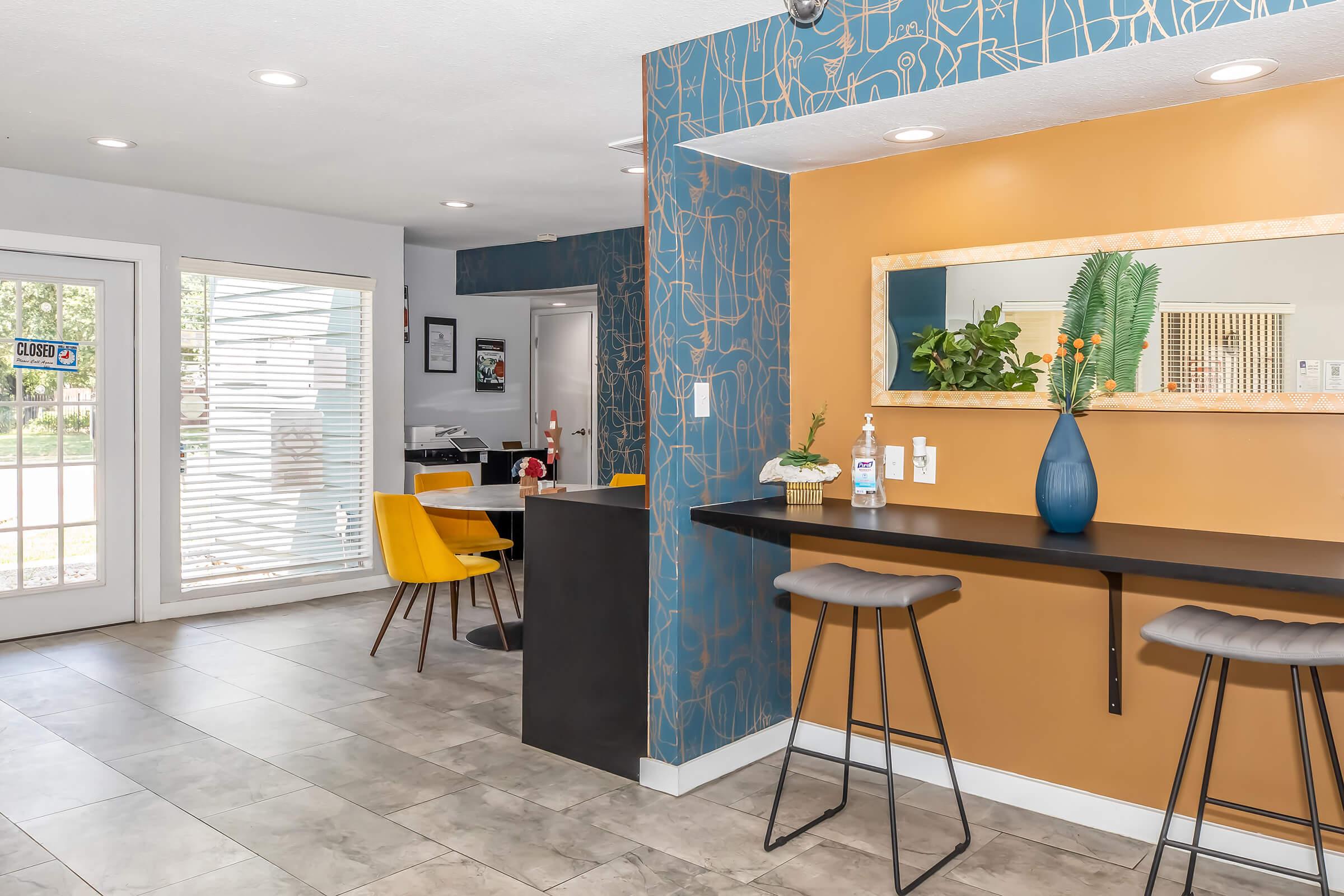 A bright and modern interior featuring a small bar with high stools and a blue vase with flowers. To the left is a sitting area with yellow chairs, and in the background, a door with blinds leads outside. The walls are adorned with a stylish blue and orange design, creating a welcoming atmosphere.
