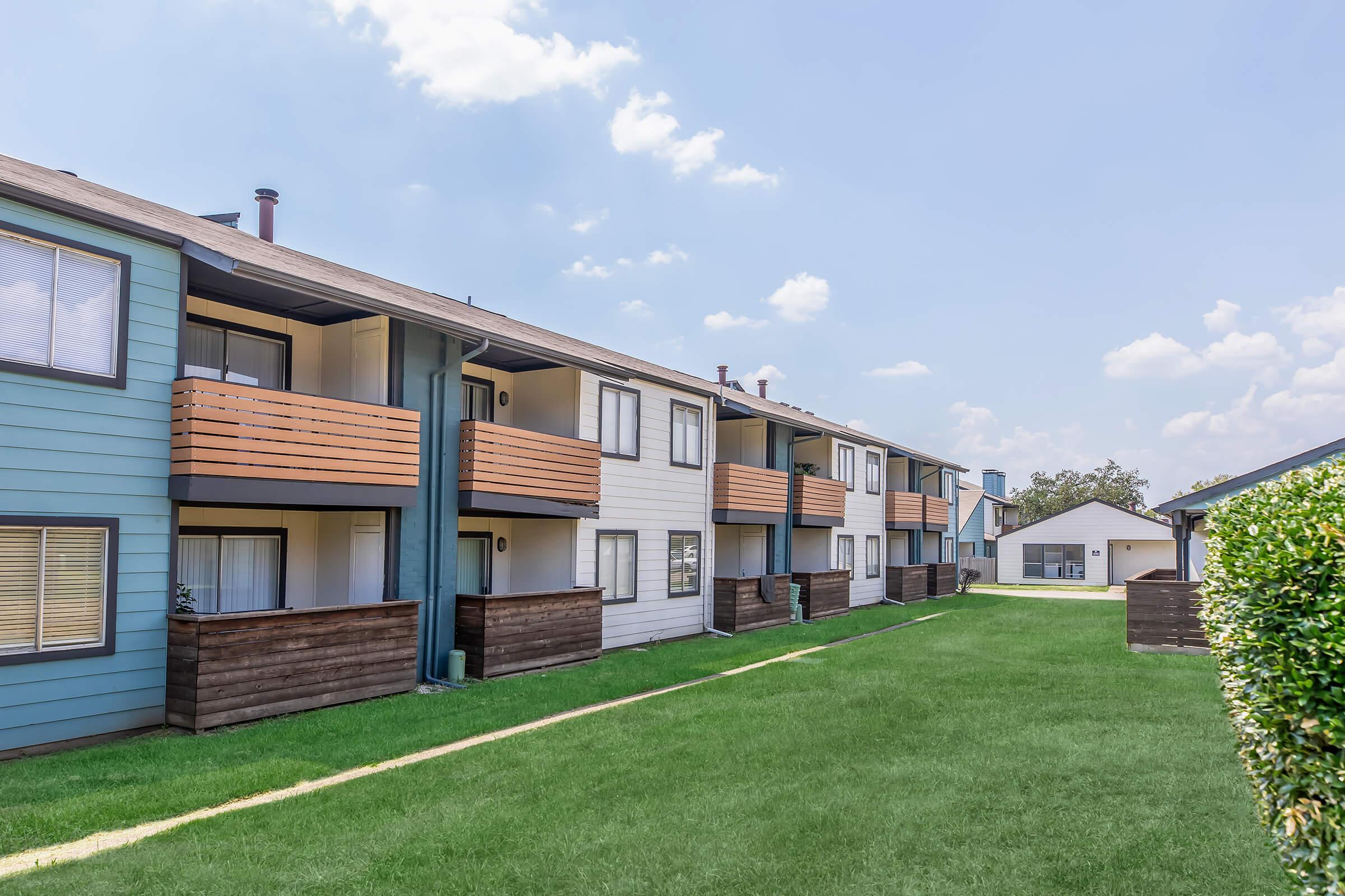 A row of modern apartment buildings with balconies, situated on a well-maintained green lawn. The buildings feature a mix of blue and white exteriors with wooden accents. In the background, a small shed is visible under a clear blue sky with scattered clouds.