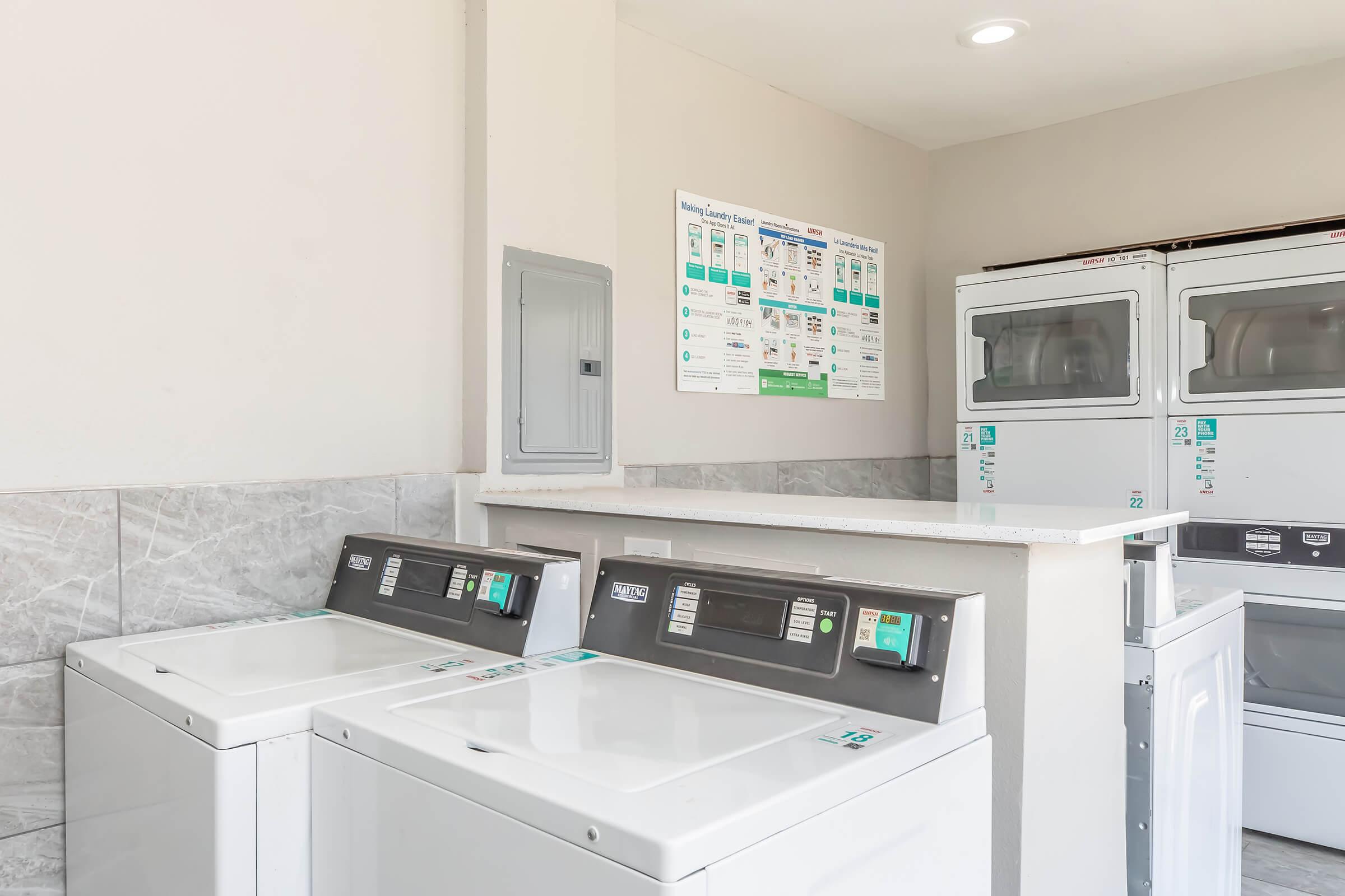 A clean laundry room featuring several washing machines and dryers. There's a countertop, a circuit breaker panel, and an informational poster on the wall displaying laundry instructions and guidelines. The environment is bright and well-maintained.