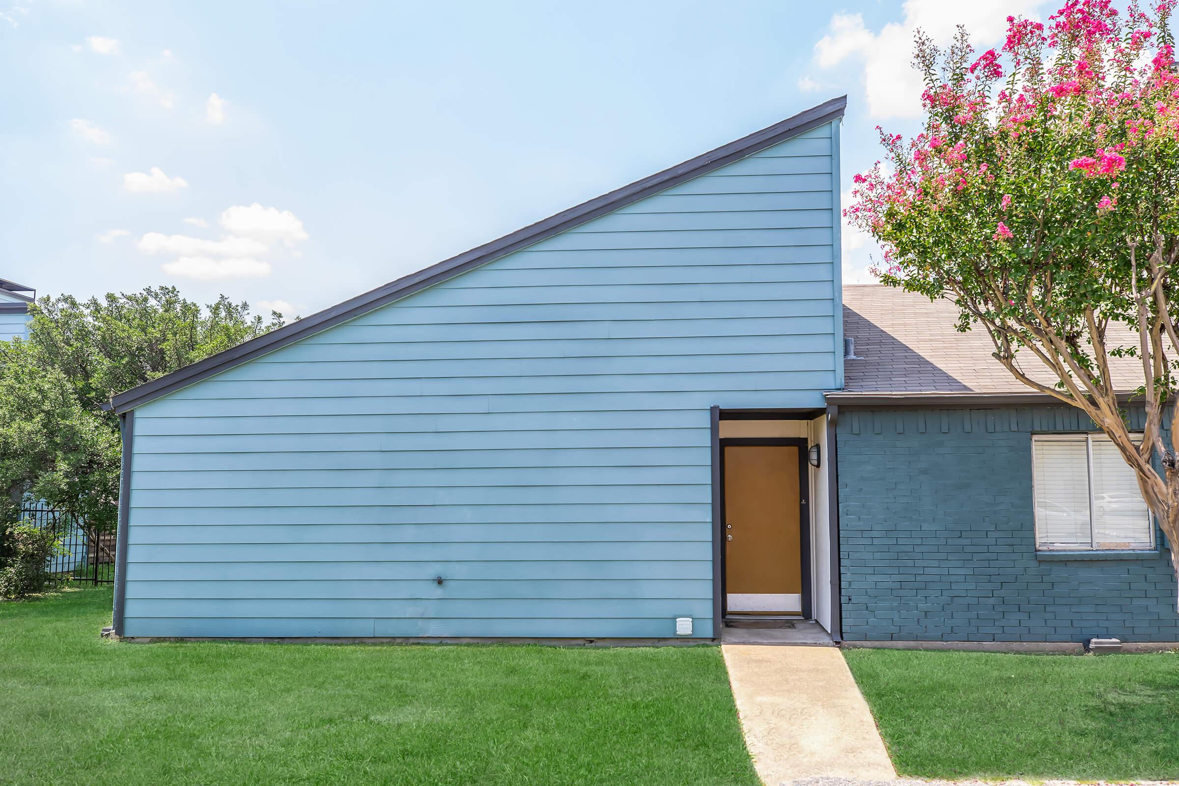 A single-story blue house with a distinct slanted roof, featuring a front door at the base of the slant. The house has a small patch of green lawn in front and is set against a clear sky with a few clouds. A flowering tree is visible nearby.
