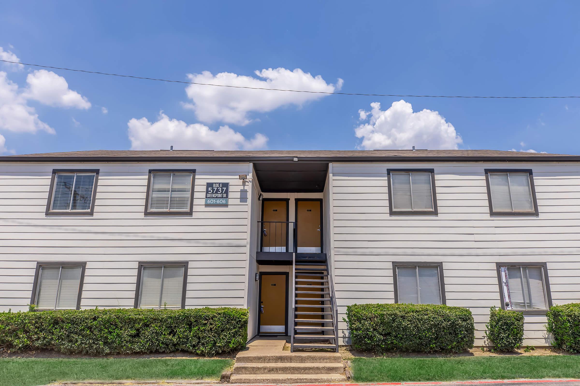 A two-story apartment building with white siding and brown accents. It features a central staircase leading to two doors on each floor. Lush green landscaping lines the entrance, and the sky above is bright blue with fluffy white clouds.