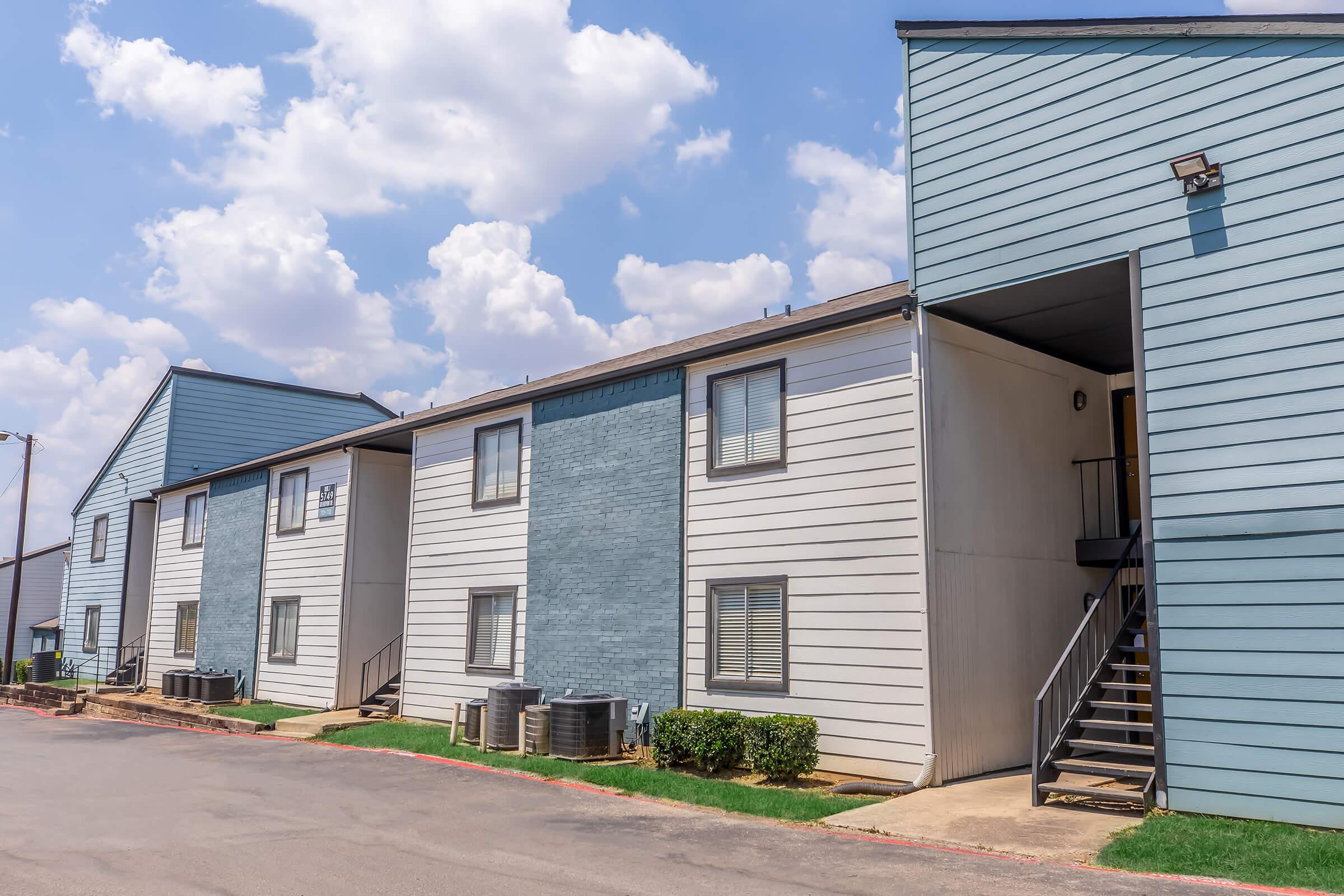Two multi-unit apartment buildings with a light blue and gray exterior. Each building has multiple windows and air conditioning units. A staircase leads to the entrance of one building. The sky is mostly cloudy, and the surrounding area is well-maintained with grass.