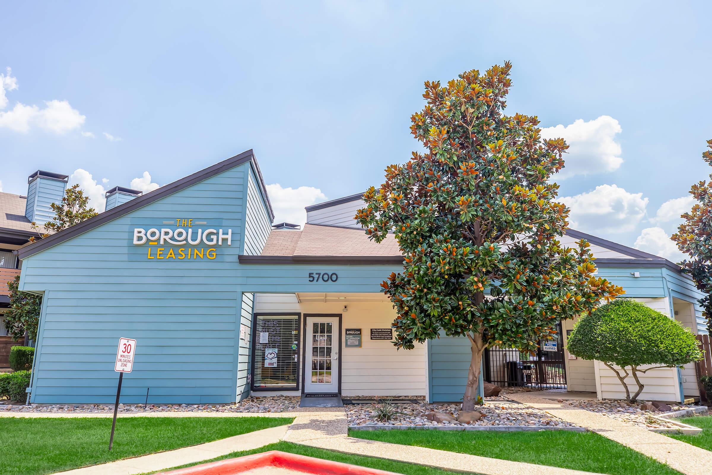 Leasing office of The Borough, a residential community, featuring a light blue exterior with yellow signage. A well-maintained tree is visible in front, and the building is surrounded by neatly landscaped grounds under a bright sky.