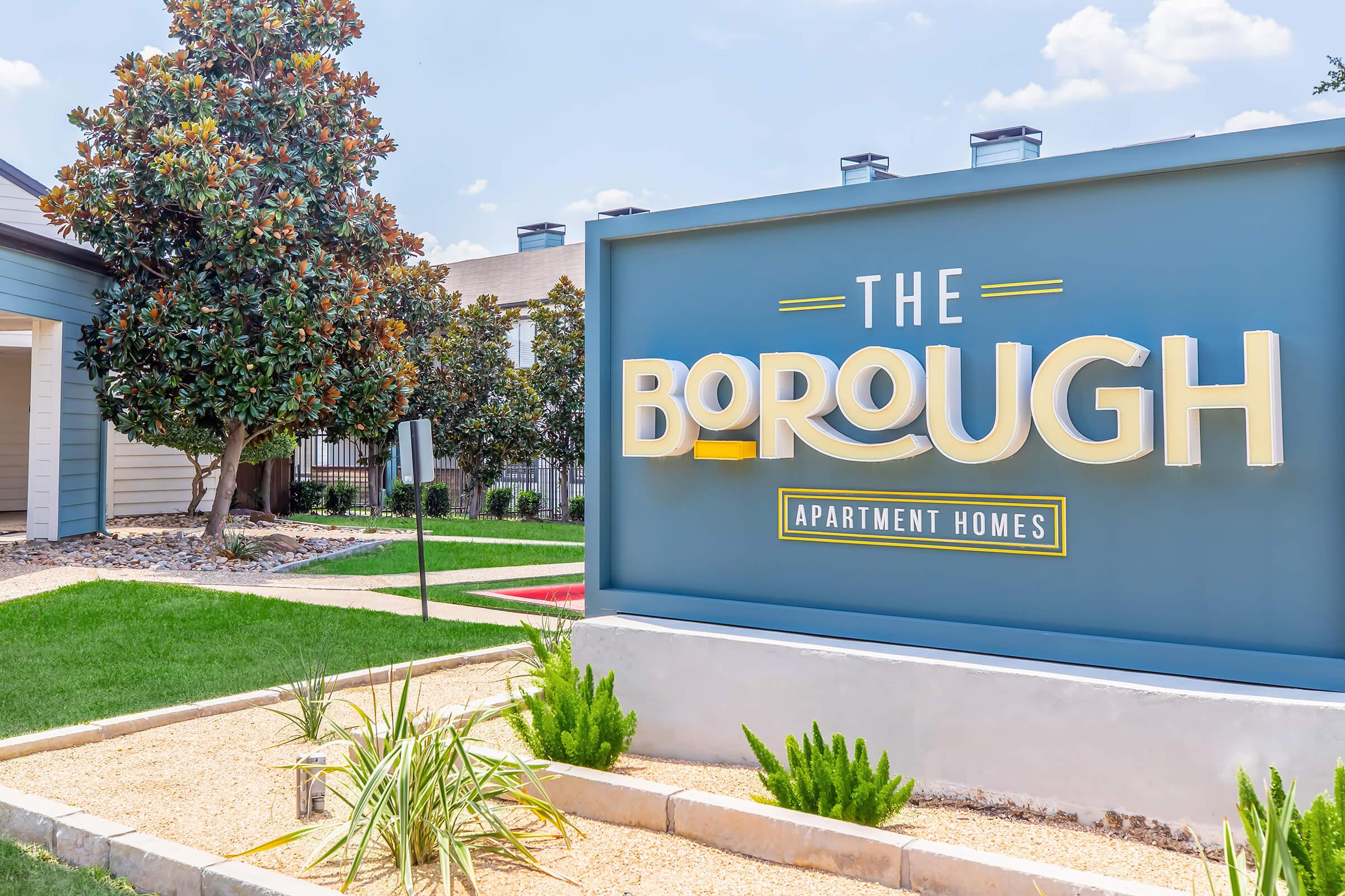 Sign for The Borough Apartment Homes, featuring bold yellow lettering against a blue background. Surrounding landscaping includes green grass and decorative plants, with a magnolia tree in the background under a clear blue sky.