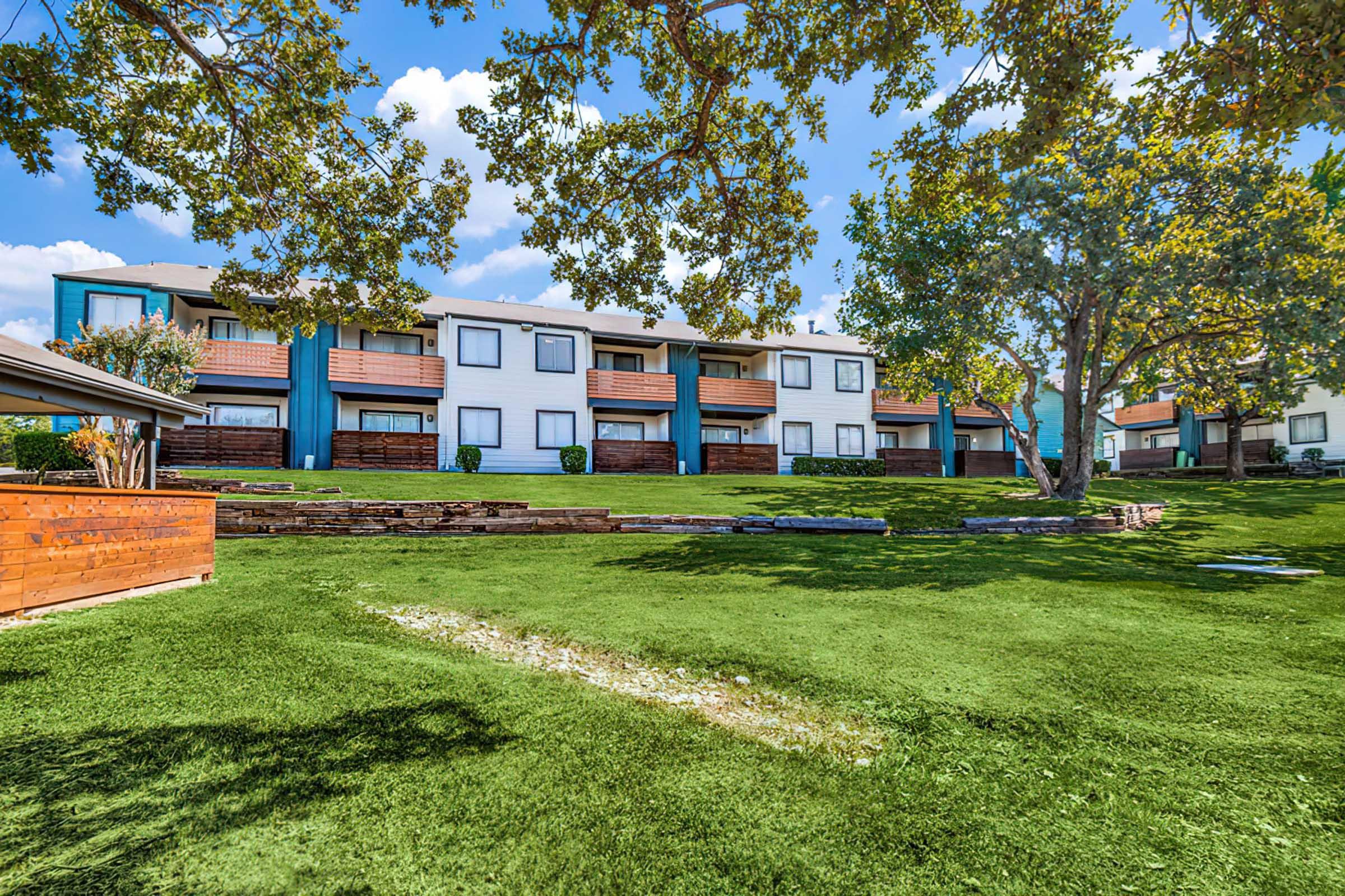 A well-maintained green lawn in front of two-story apartment buildings with balconies. The buildings are surrounded by trees and landscaping, featuring a modern design with a blue and white color scheme. Fluffy white clouds are visible in a bright blue sky.