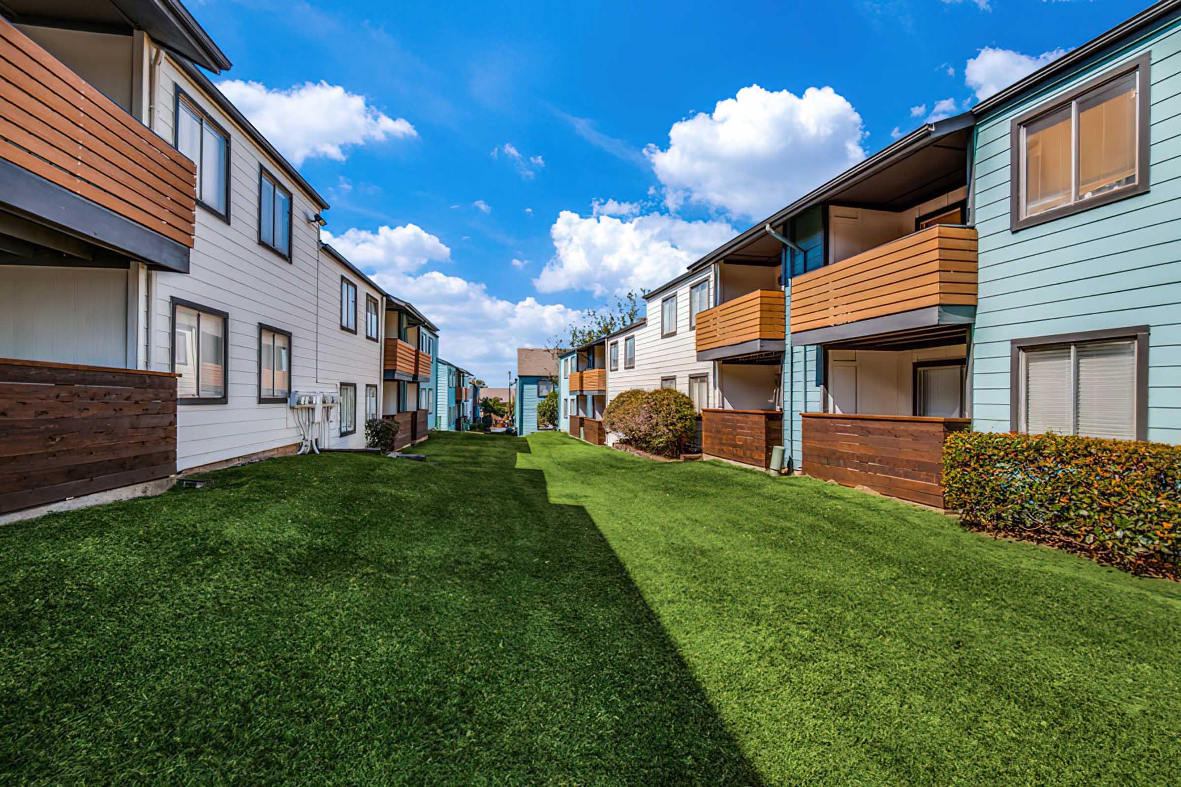 A view of a landscaped pathway between two sets of apartment buildings. The grassy area is filled with lush green turf, flanked by small shrubs and bushes. The buildings have a mix of light and dark siding with balconies, under a bright blue sky with scattered clouds.