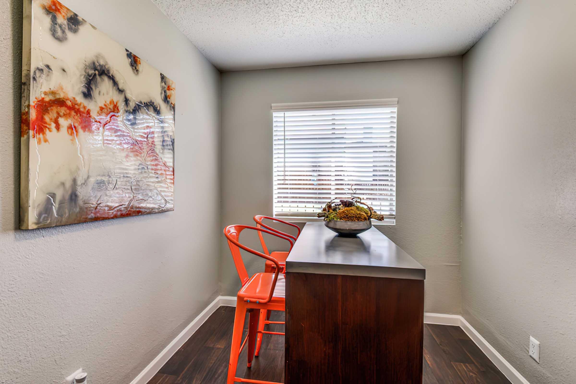 A small, modern workspace featuring a sleek wooden desk and two bright orange chairs. A large abstract painting with red, white, and blue tones is hung on the wall. A window with white blinds allows natural light to enter, illuminating the simple, stylish decor.