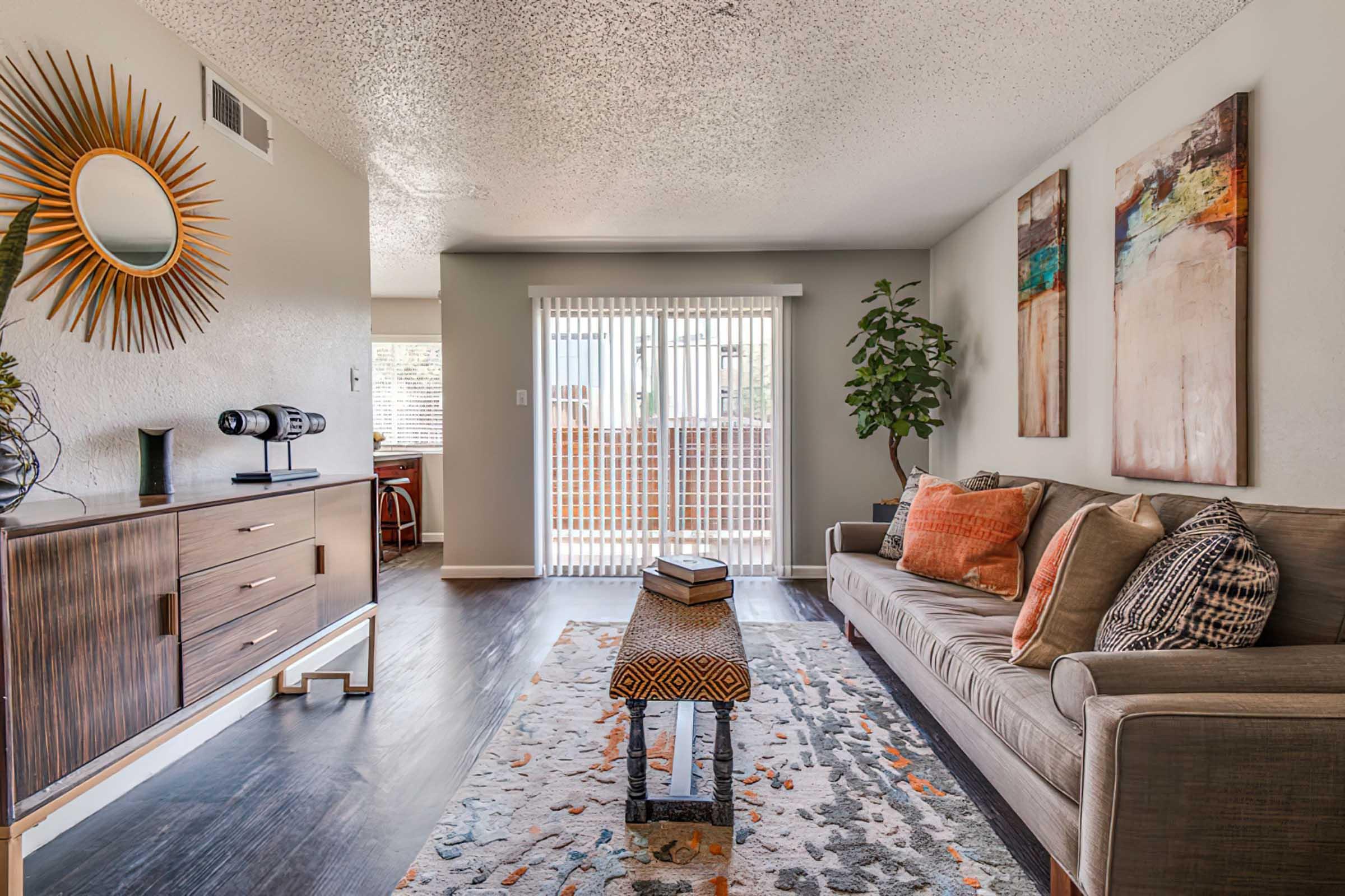 A cozy living room featuring a modern sofa adorned with decorative pillows, a stylish area rug, a wooden coffee table with stacked books, and a decorative console. Large windows provide natural light, and abstract artwork hangs on the walls. A plant adds a touch of greenery to the warm, inviting atmosphere.