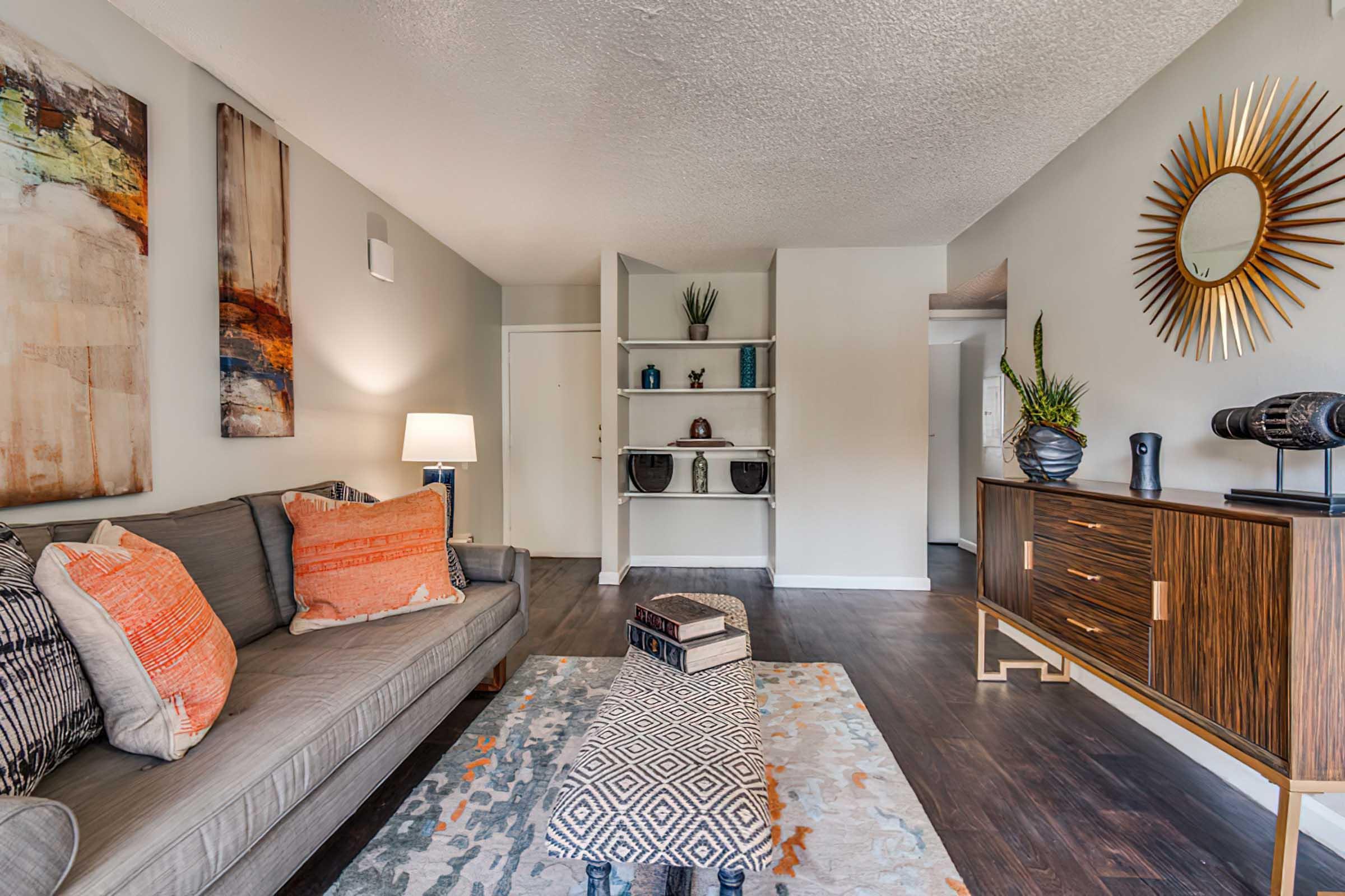 A modern living room featuring a gray sofa adorned with patterned cushions, an area rug with geometric designs, and a stylish wooden sideboard. The walls are decorated with abstract art, and there’s a round mirror on one wall. Soft lighting from a lamp creates a cozy atmosphere.