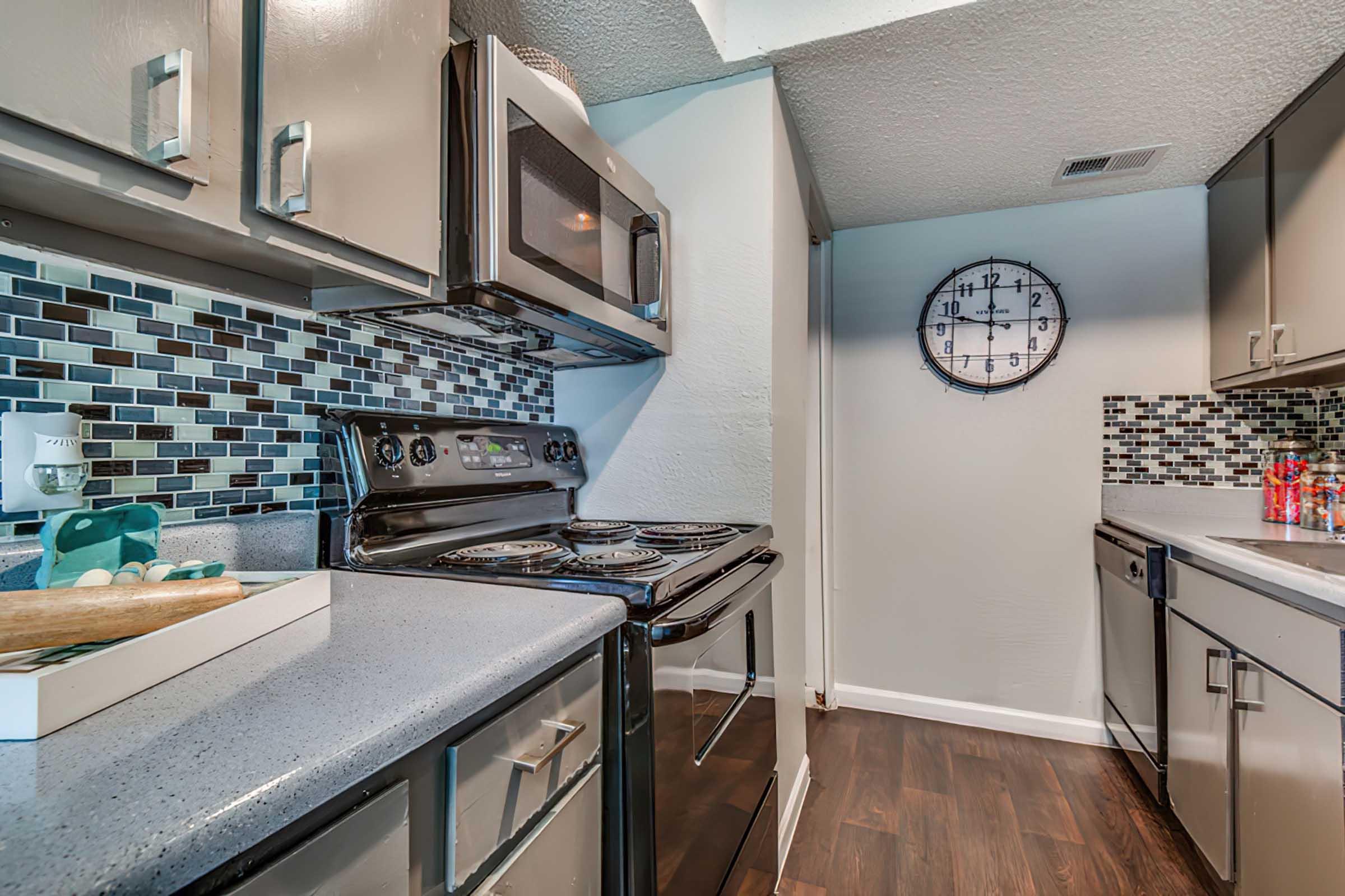 Modern kitchen featuring stainless steel appliances including a microwave and stove, complemented by mosaic tile backsplash and a round wall clock. The countertops are dark, with a light-colored cabinet layout. The flooring is dark wood, creating a warm atmosphere. A tray with decorative items is visible.