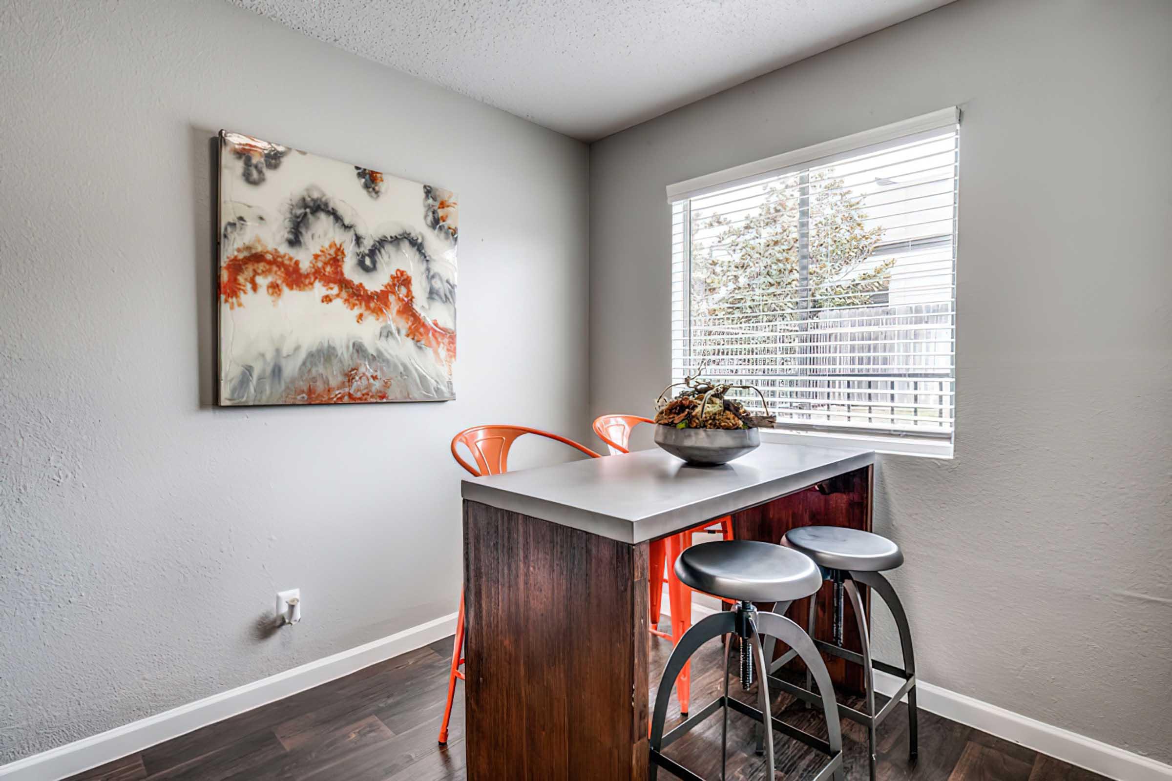 A modern dining area featuring a sleek bar table with two black stools and orange chairs. The table is adorned with a decorative bowl. A large abstract painting with orange and gray tones hangs on the wall opposite a window that lets in natural light, illuminating the space.