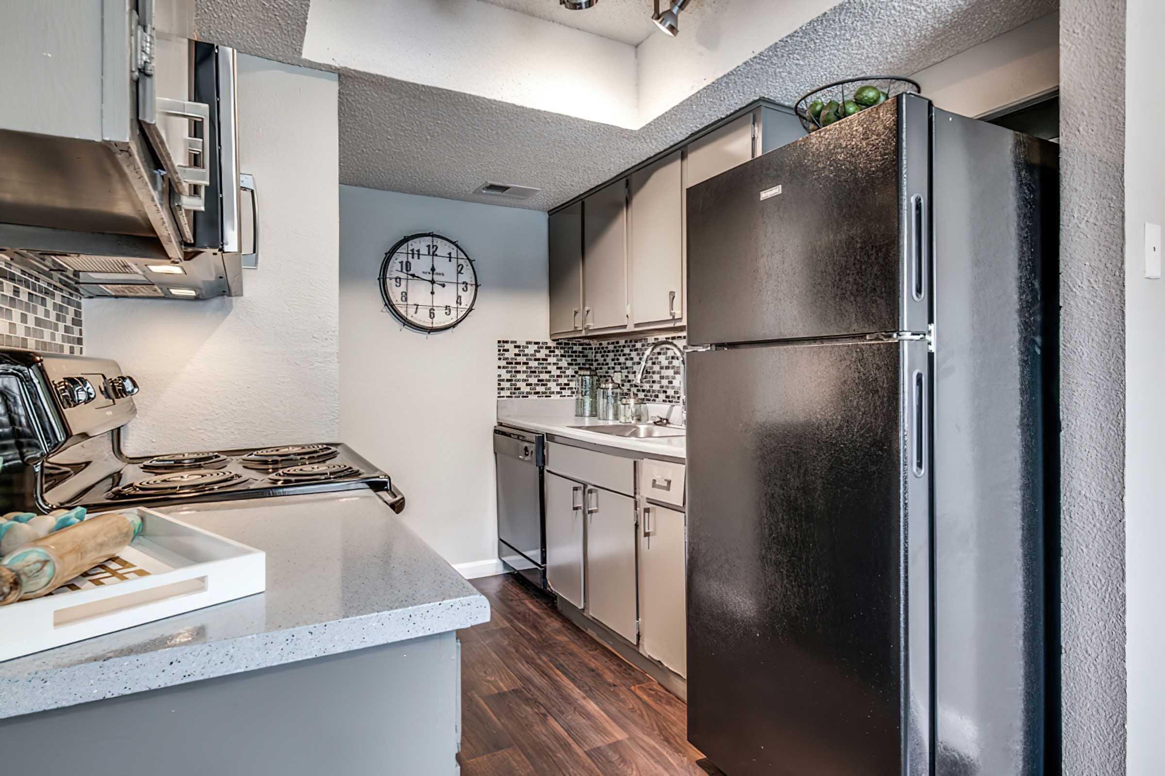 Modern kitchen featuring stainless steel appliances, a black refrigerator, and a tiled backsplash. A large clock is mounted on the wall, and the space has dark wood flooring. Countertops are light-colored with a simple tray on one side. Overall, the design is sleek and contemporary.
