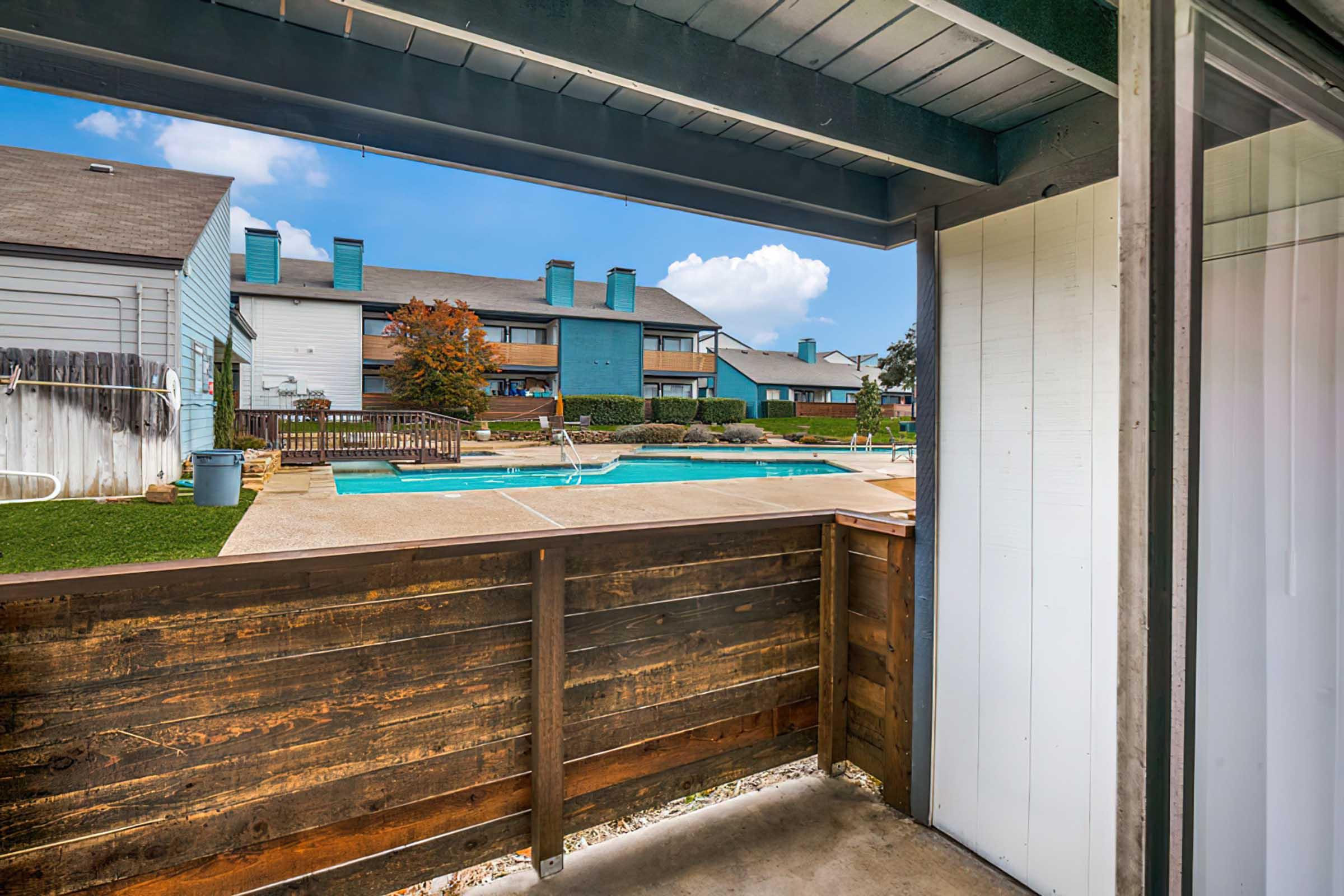 View from a balcony overlooking a swimming pool area, surrounded by landscaped grounds and residential buildings. The sky is clear with a few clouds, and the setting appears inviting and well-maintained.