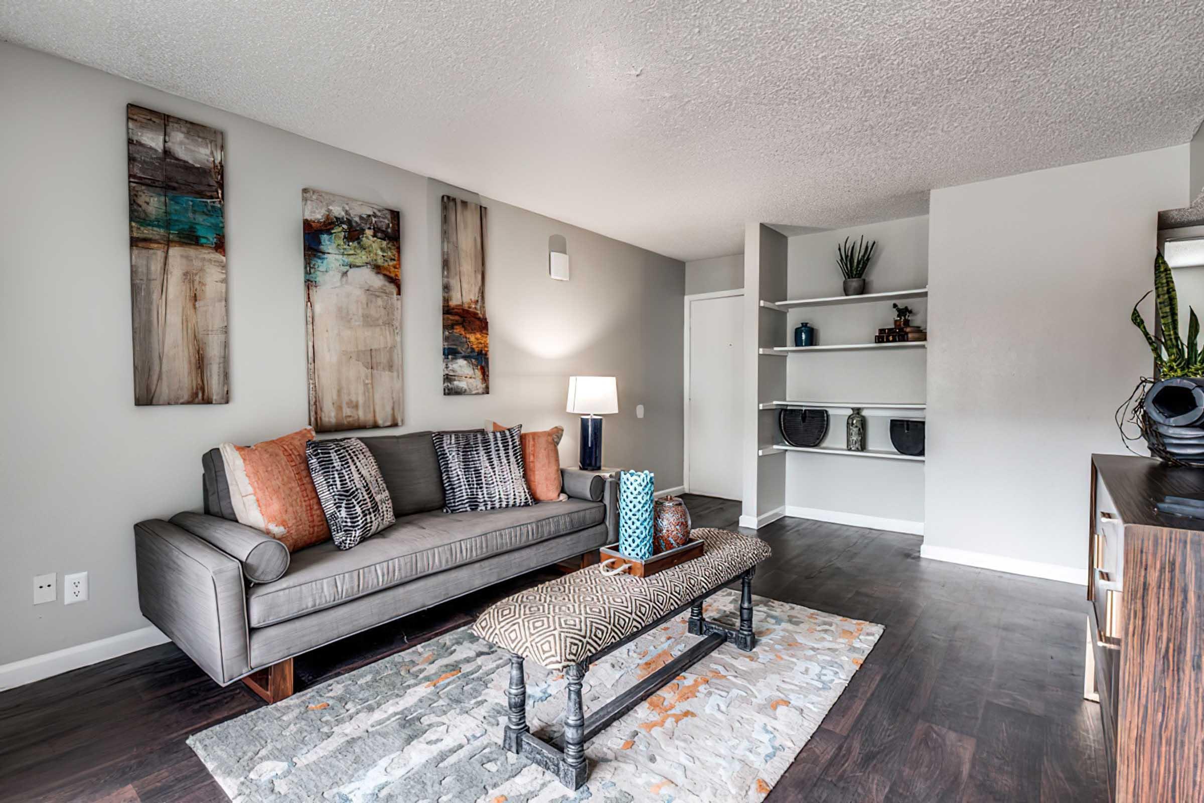 A modern living room featuring a grey sofa with colorful accent pillows, a patterned coffee table, and decorative items. Wall art hangs above the sofa, and shelves with vases are displayed on the right. The space is bright with neutral-colored walls and a warm wooden floor.