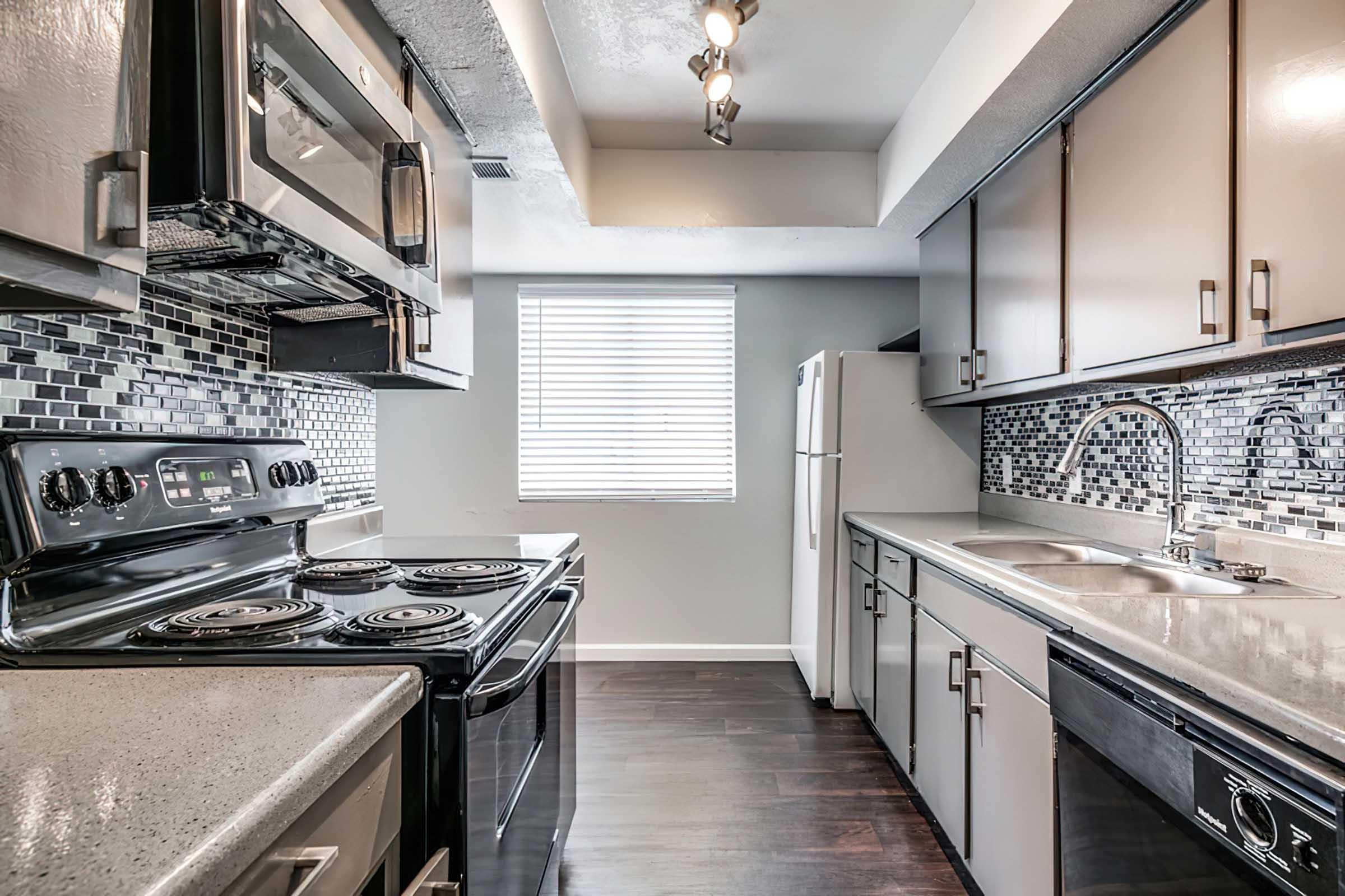 Modern kitchen with stainless steel appliances, including an oven and refrigerator. The countertops are light-colored with a sleek finish, and the backsplash features black and gray tiles. A window above the sink allows natural light, and there are dark wood cabinets providing storage space.