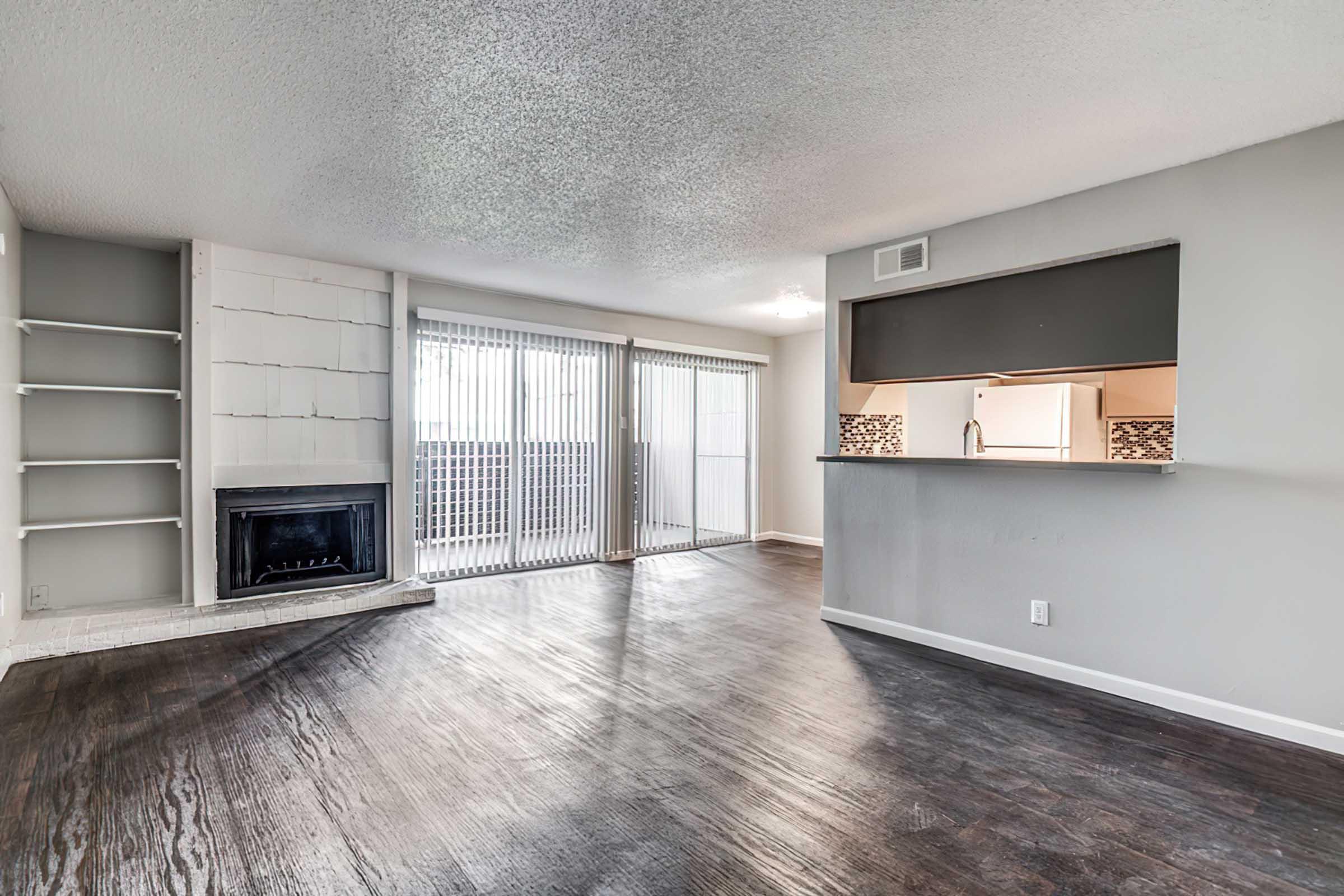 Spacious interior of an apartment showcasing a living area with a large window and sliding glass doors leading to a balcony. Features include a fireplace and an open kitchen with a bar area, all set against a backdrop of light-colored walls and dark wood flooring.