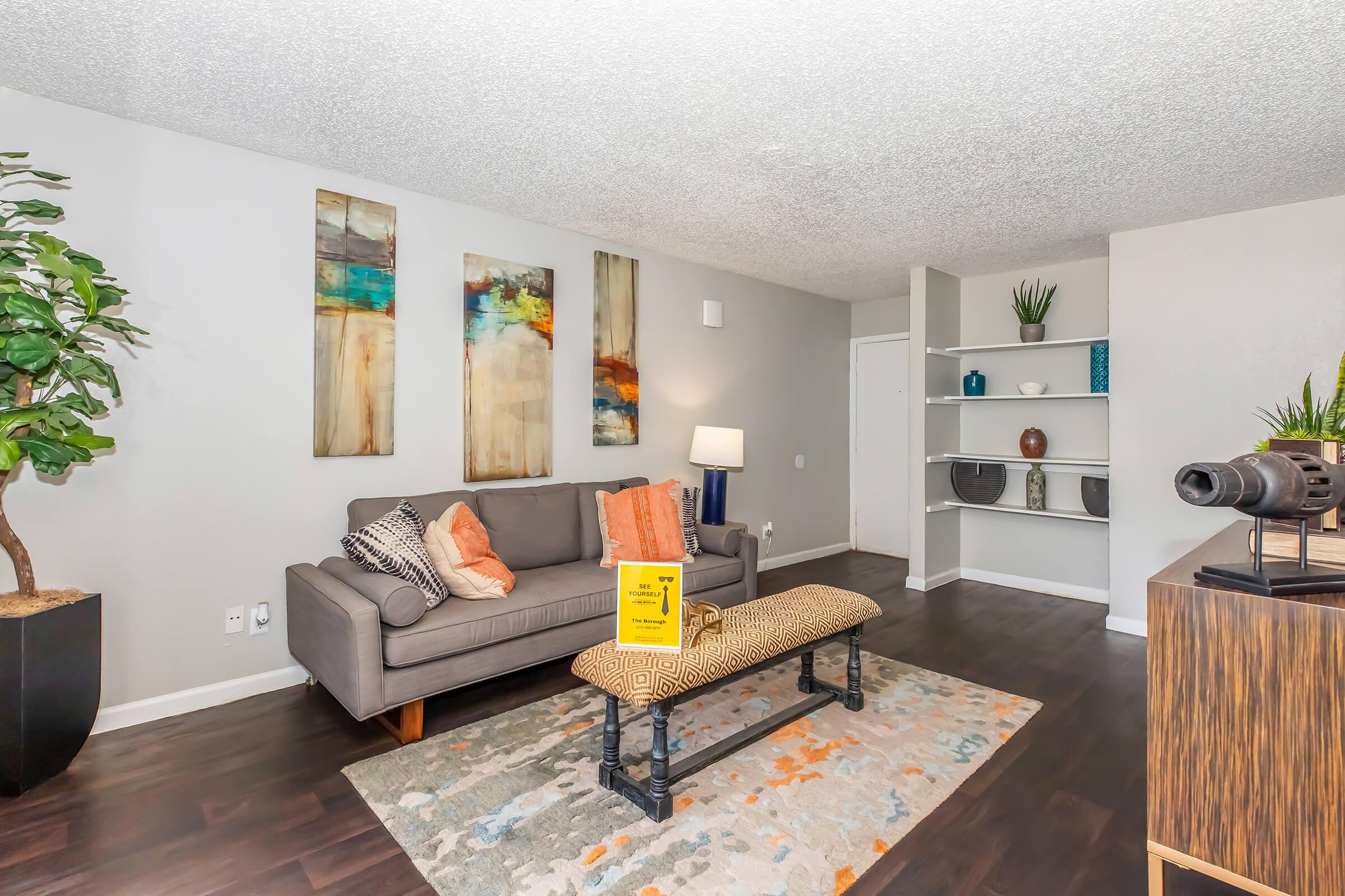 A modern living room featuring a grey couch with colorful throw pillows, an abstract wall art arrangement, a small table with a decorative book, and a patterned area rug. There is a floor lamp beside the couch, a plant in a pot, and open shelving with decorative items on one side. The walls are light-colored, creating a bright atmosphere.