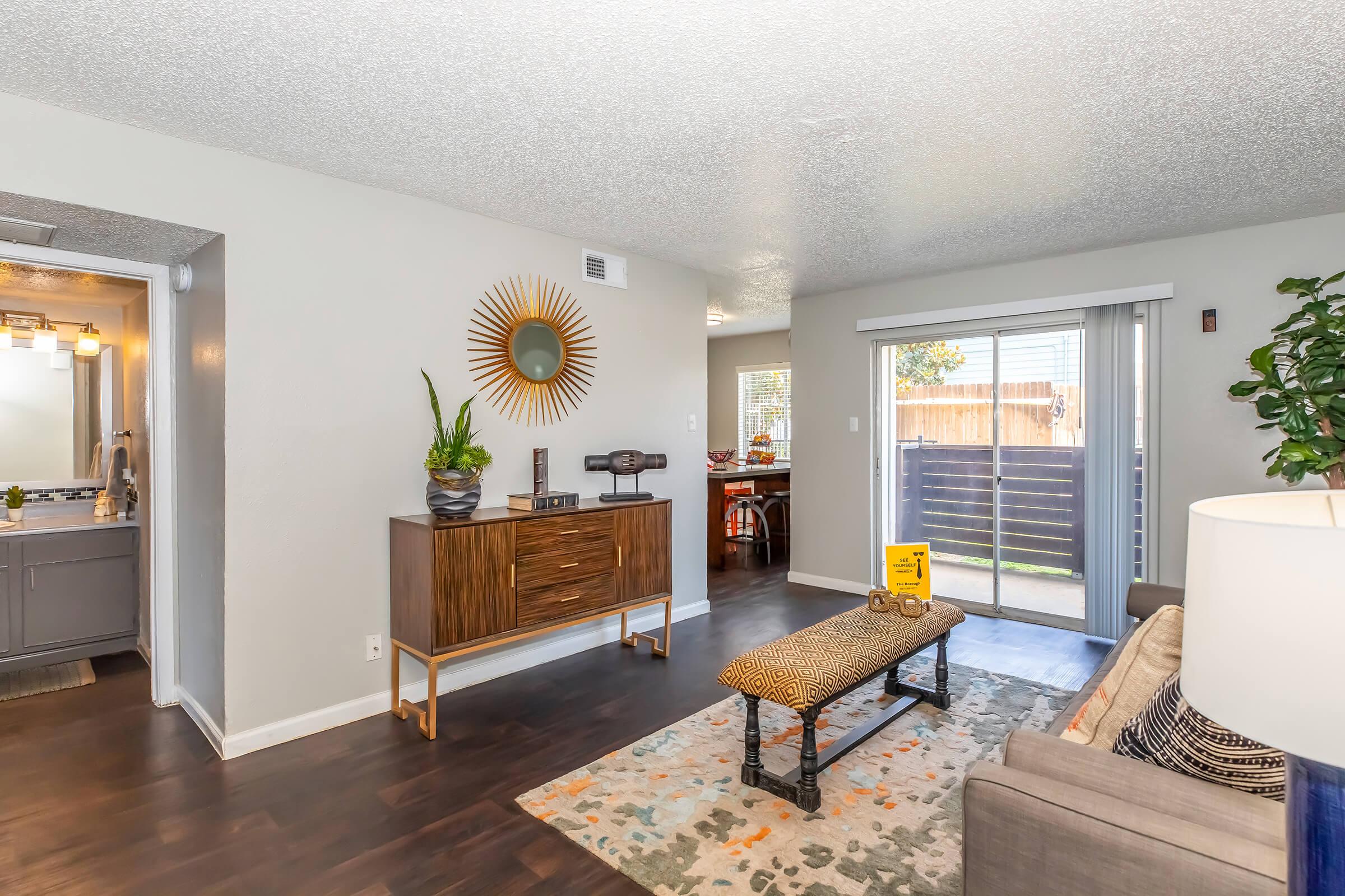 Modern living room featuring a stylish gray sofa, a decorative coffee table with a patterned runner, and a mid-century modern sideboard with a round mirror above it. Natural light floods in through sliding glass doors that lead to an outdoor area, with a glimpse of a dining space in the background.