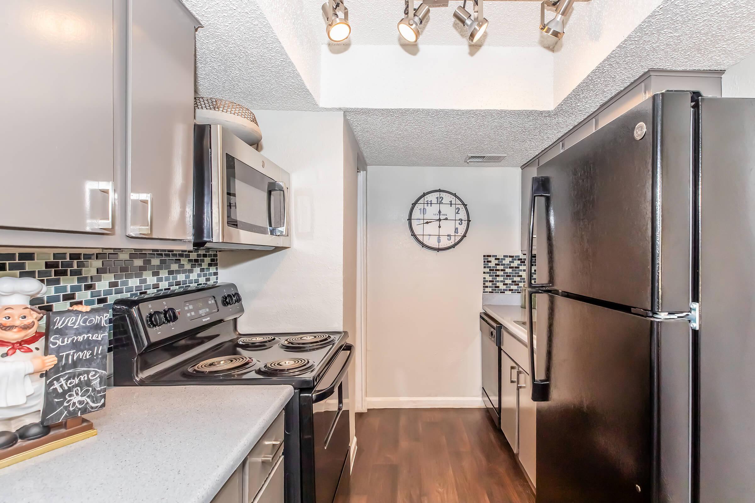 A modern kitchen featuring stainless steel appliances, including a microwave, stove, and refrigerator. The cabinets are a sleek gray, with a contrasting dark countertop. Decorative elements include a wall clock and a chef figurine on the counter, while the backsplash displays a mosaic of blue and black tiles.
