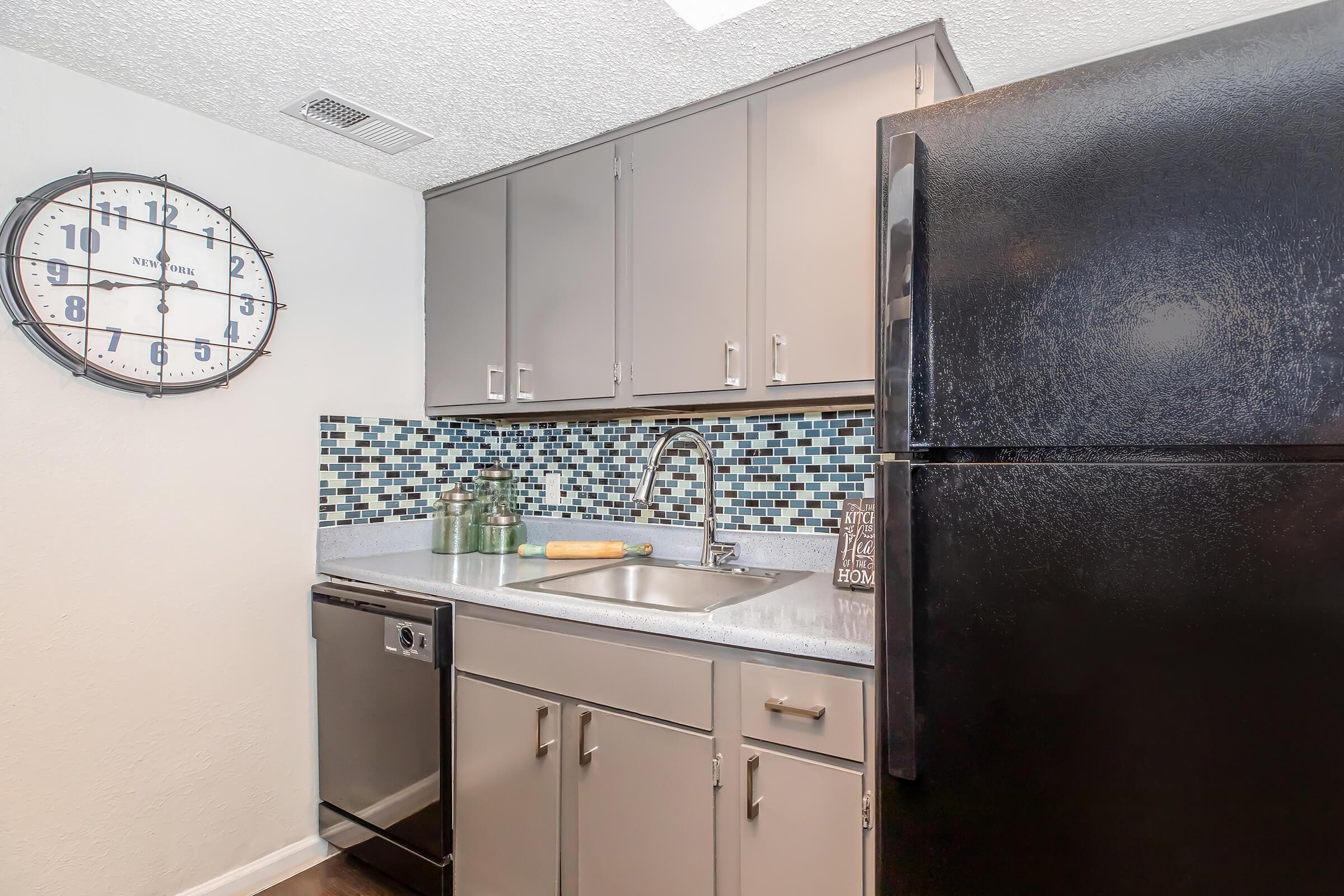 A modern kitchen featuring gray cabinetry, a stainless steel dishwasher, and a black refrigerator. The backsplash is adorned with blue mosaic tiles, and a round wall clock is displayed above the sink, which has a contemporary faucet. A rolling pin and decorative items are placed on the countertop.