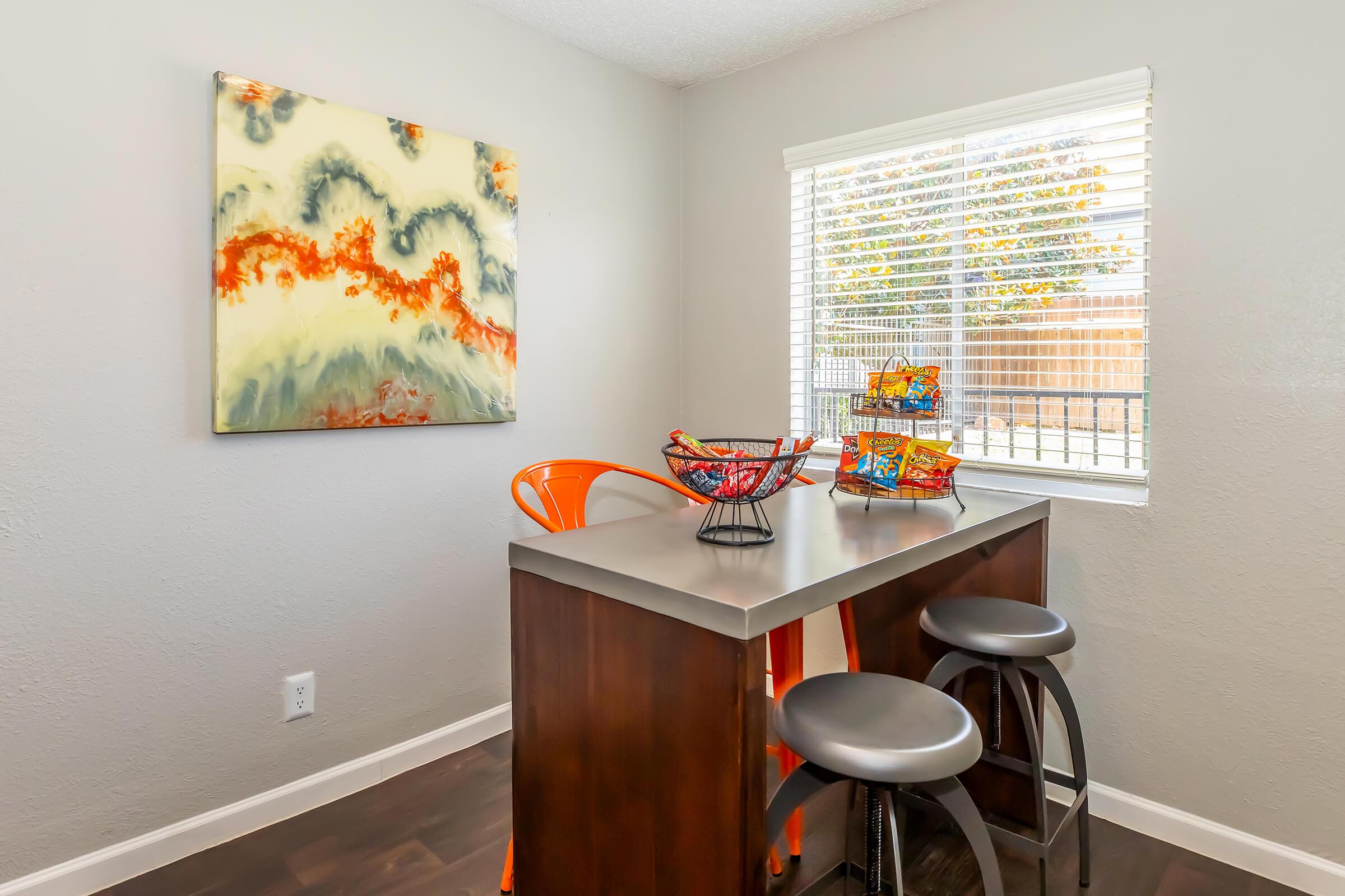 A modern dining area featuring a small bar-style table with two black stools. On the table, there is a colorful fruit bowl and a decorative basket. A vibrant abstract painting hangs on the wall, complementing the bright orange chair nearby. Natural light streams in through the window, illuminating the space.
