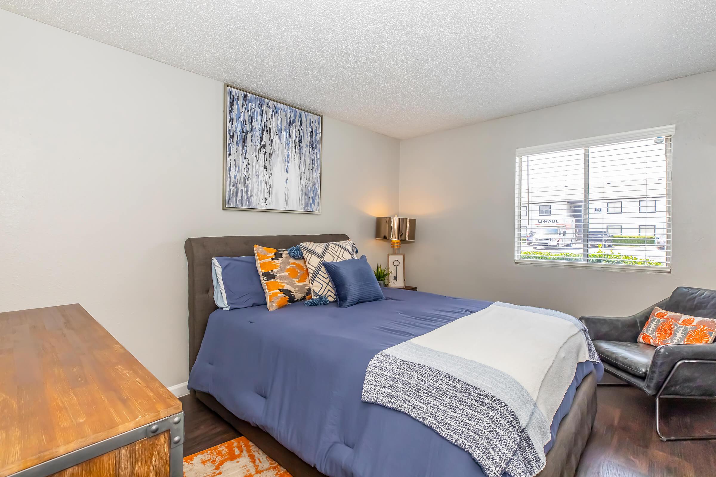 A cozy bedroom featuring a queen-sized bed with blue bedding and decorative pillows. There's a modern piece of artwork on the wall, a wooden nightstand with a lamp, and a black leather armchair with orange accents. Natural light streams in through a window with blinds.