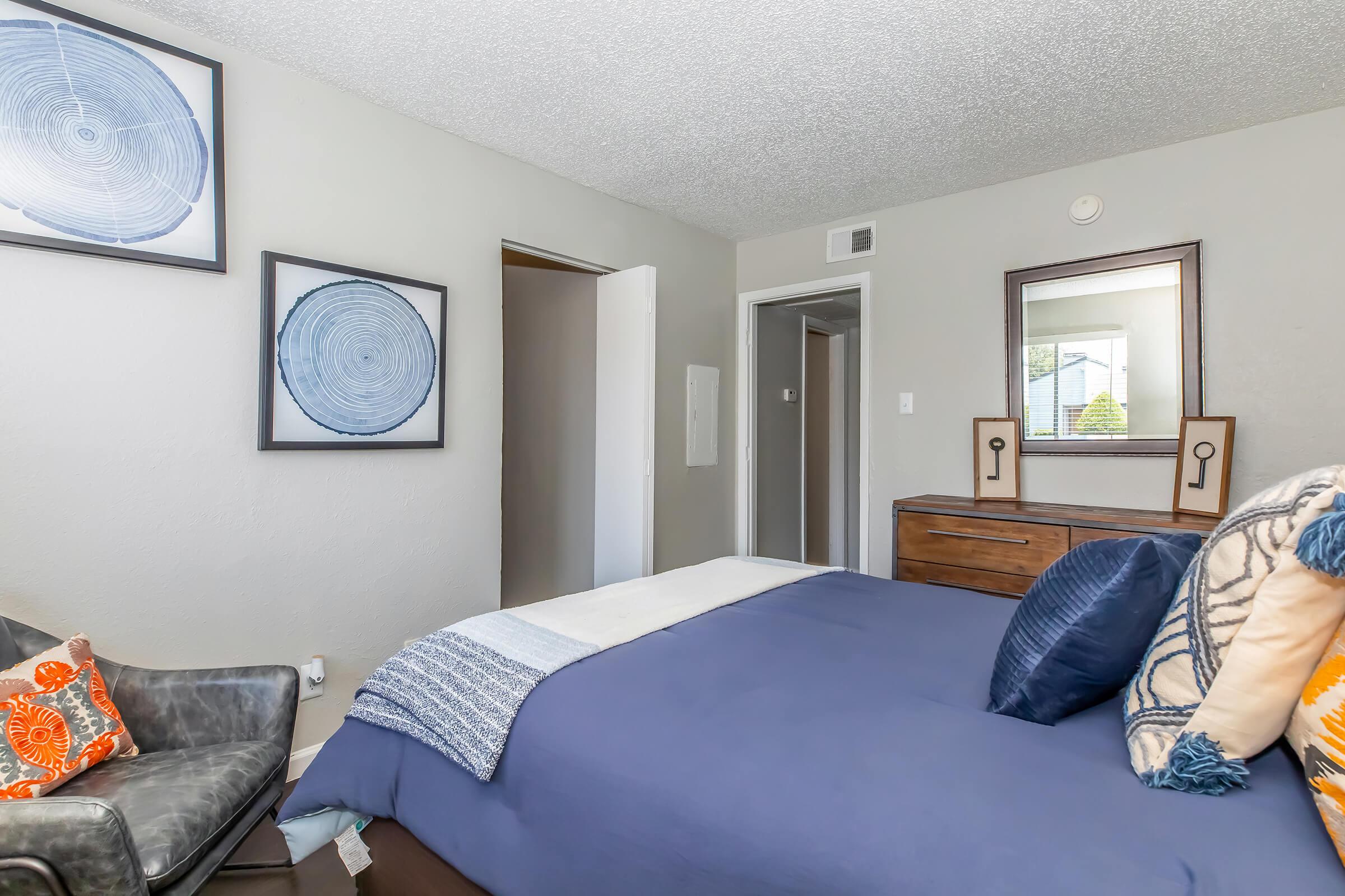 A cozy bedroom featuring a blue bedspread with decorative pillows, a wooden dresser, and framed circular art on the walls. There is a chair in the foreground, and a door leading to another room in the background. Natural light illuminates the space, creating a warm and inviting atmosphere.