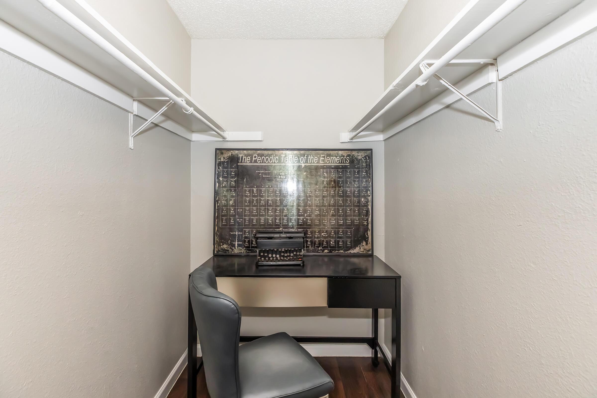 A small, organized closet featuring a dark wall-mounted panel with elements displayed in a periodic table format. A black desk and chair are positioned in front of the panel, creating a compact workspace. The walls are painted in a light color, and the floor is dark wood.