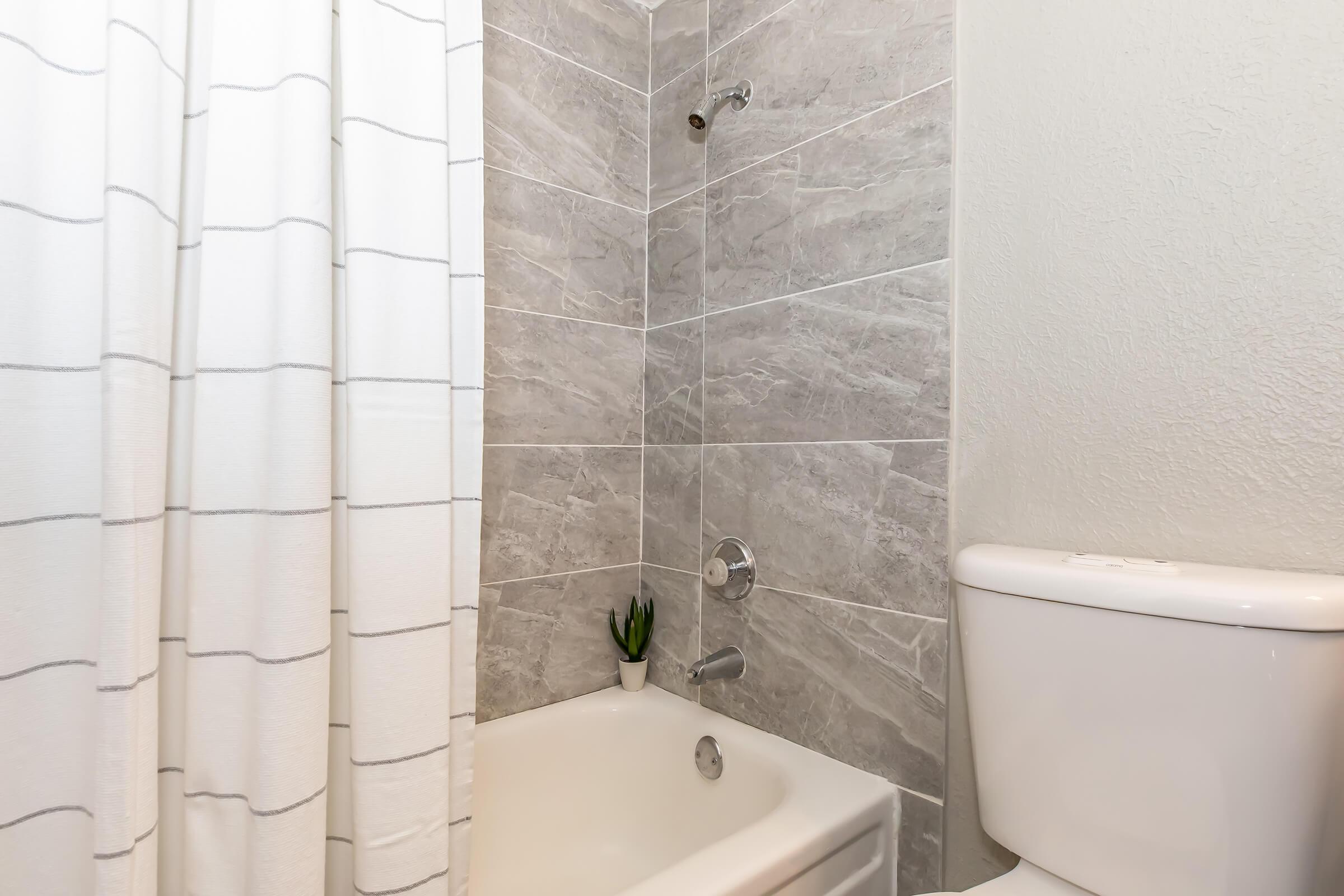 A clean, modern bathroom featuring a bathtub with a curved shower rod and a white curtain. The walls are tiled in gray with light-colored grout, and there is a small potted plant on the edge of the bathtub. A white toilet is positioned next to the tub, creating a minimalist and inviting space.