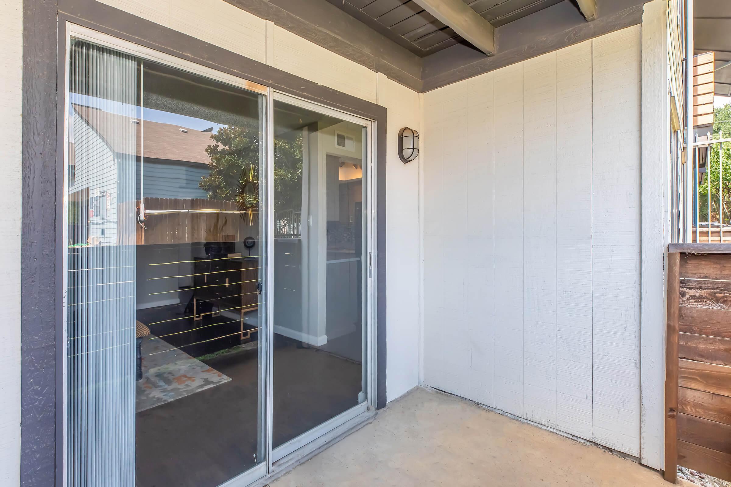 A porch area featuring a sliding glass door that leads into an interior space. The wall has a light fixture, and the floor is a concrete slab. Outside, there are glimpses of greenery and neighboring buildings. The overall setting appears to be a residential area, contributing to a cozy atmosphere.
