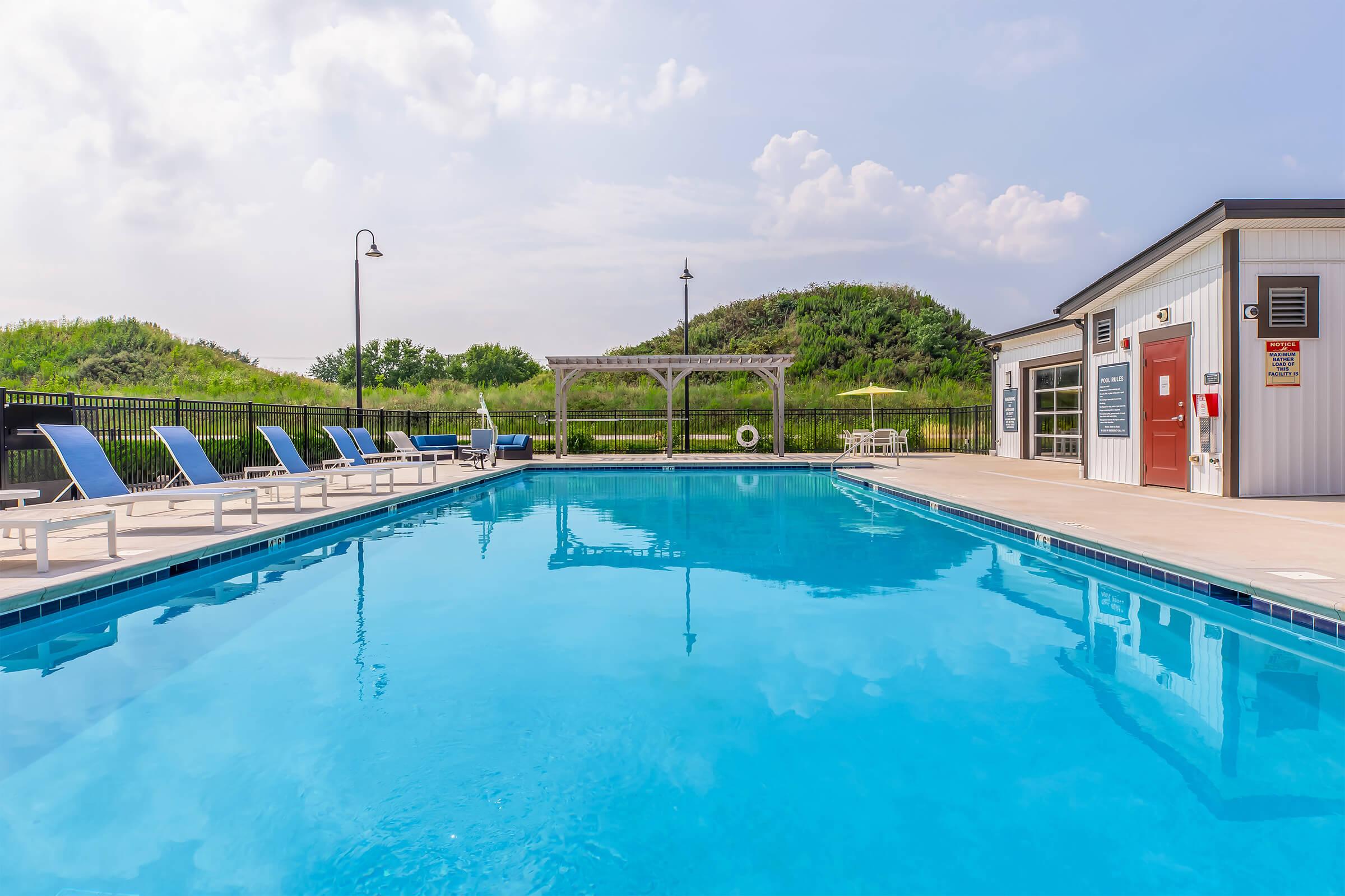 A clear blue swimming pool surrounded by lounge chairs. Two outdoor light fixtures are visible. In the background, there are grassy hills and a small pavilion beside the pool. The sky is partly cloudy, creating a serene and inviting atmosphere.