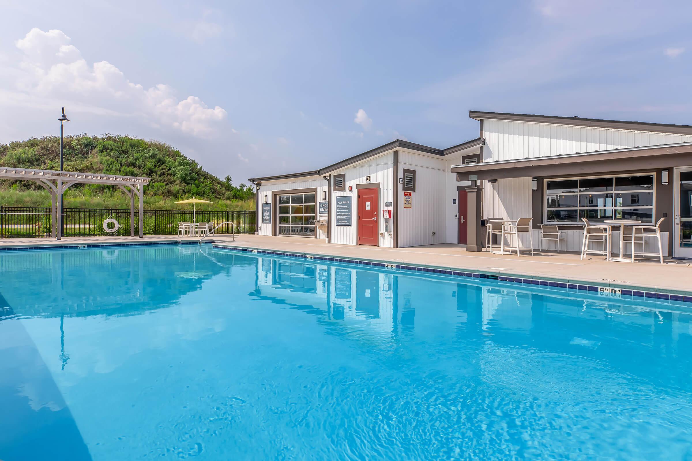 A clear blue swimming pool surrounded by a deck and lounge chairs, with a modern building in the background. The building features large windows, seating areas, and a pergola. Lush green hills are visible behind the pool under a partly cloudy sky.