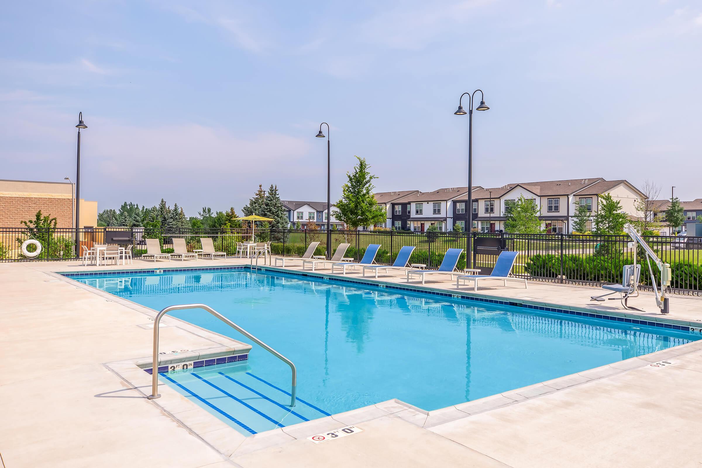 A sunny outdoor pool area featuring clear blue water, surrounded by lounge chairs. In the background, there are modern apartment buildings with trees and greenery nearby. The pool has a ladder for entry and is enclosed by a fence, with lampposts adding to the ambiance.
