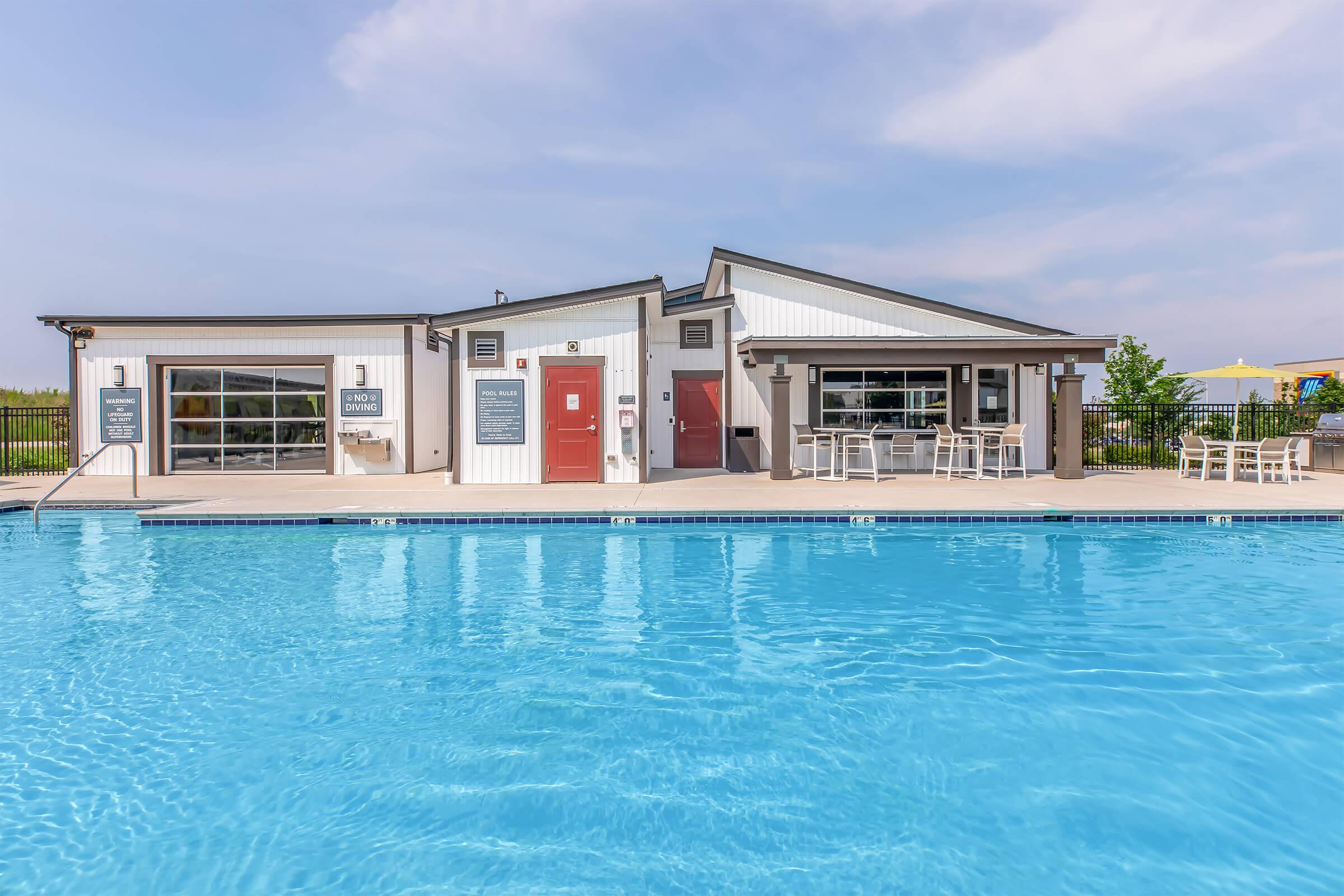 A clear blue swimming pool in the foreground, with a modern amenities building in the background. The building features large windows, a covered patio area with tables and chairs, and a sign indicating it's a community facility. The sky is partly cloudy, and there's a vibrant atmosphere.