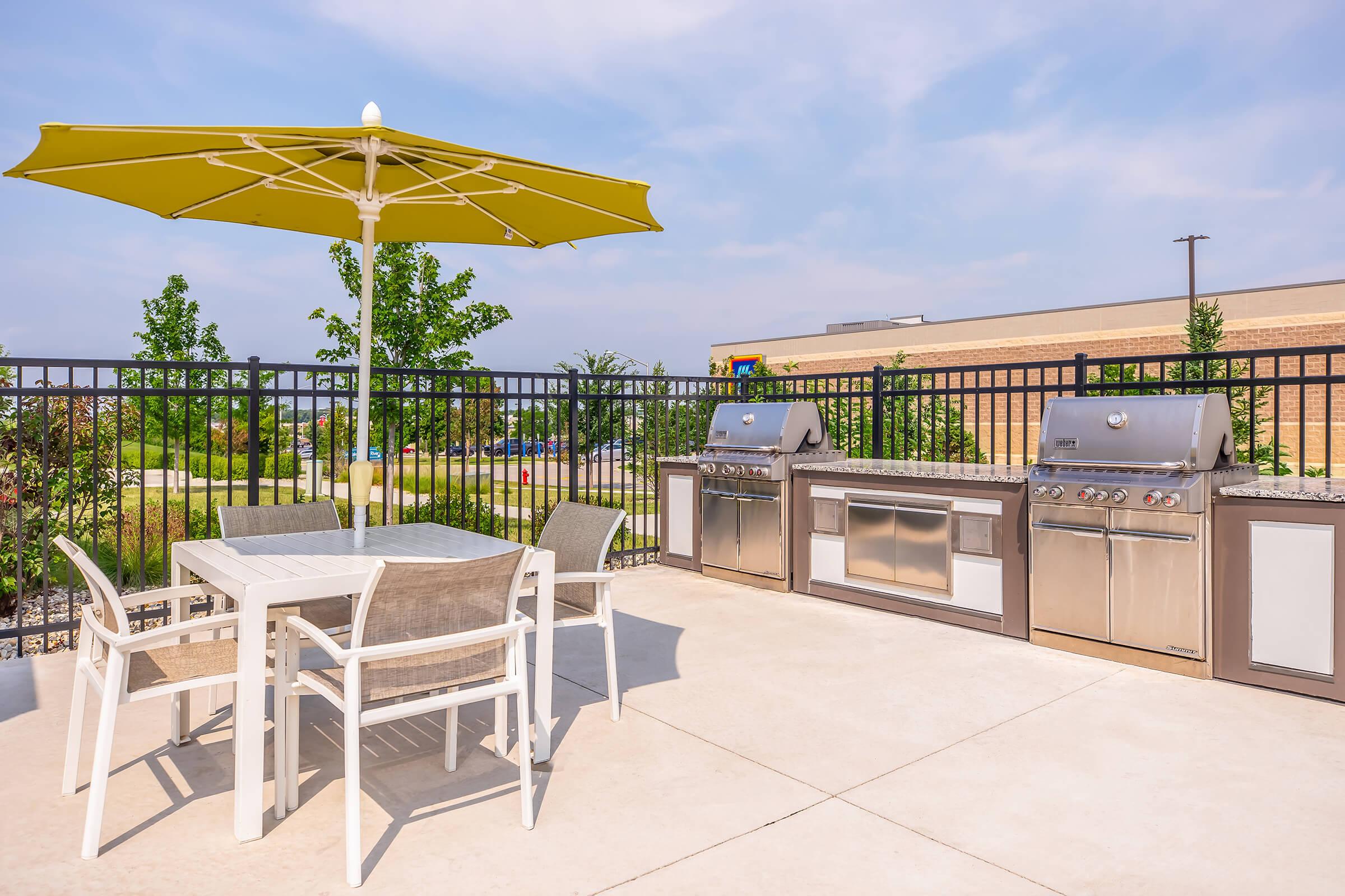 Outdoor patio area featuring a table with four chairs and a large yellow umbrella. A row of stainless steel grills is along a black fence, surrounded by green trees and a clear blue sky. Ideal setting for barbecues and gatherings.