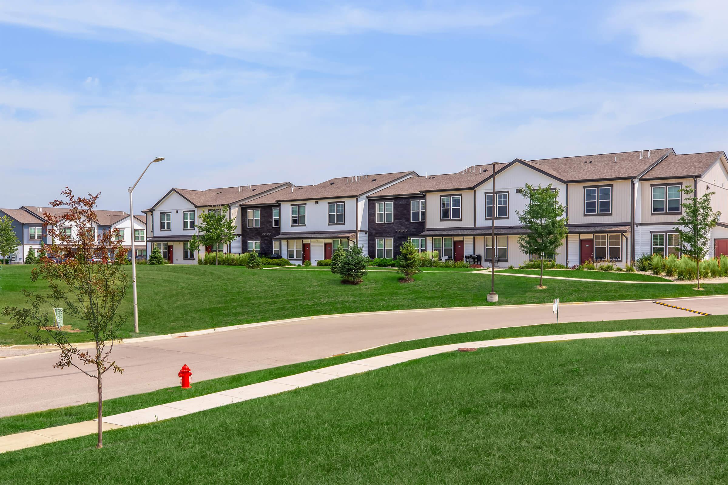 Row of modern townhouses with green lawns and trees in a suburban setting. The road curves around the foreground, leading to the houses. The sky is partly cloudy, and the overall atmosphere is peaceful and inviting.