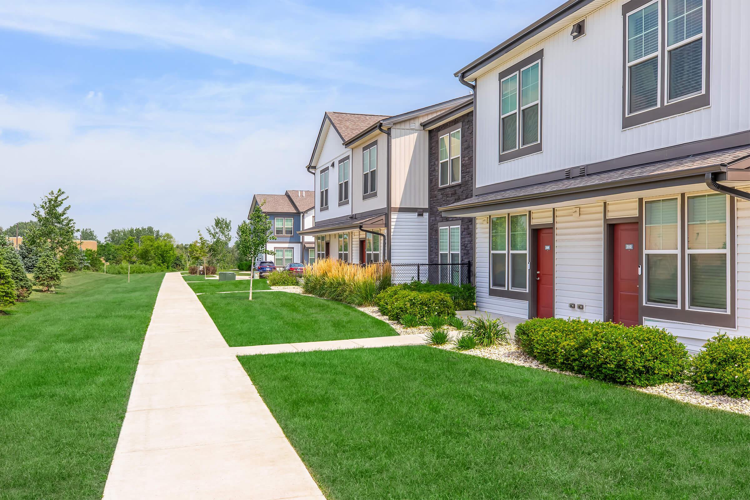 A well-maintained residential complex featuring two-story townhouses with grassy lawns, landscaped bushes, and a concrete pathway. The atmosphere is bright and sunny, with a clear blue sky and trees in the background. The layout showcases a peaceful community setting.