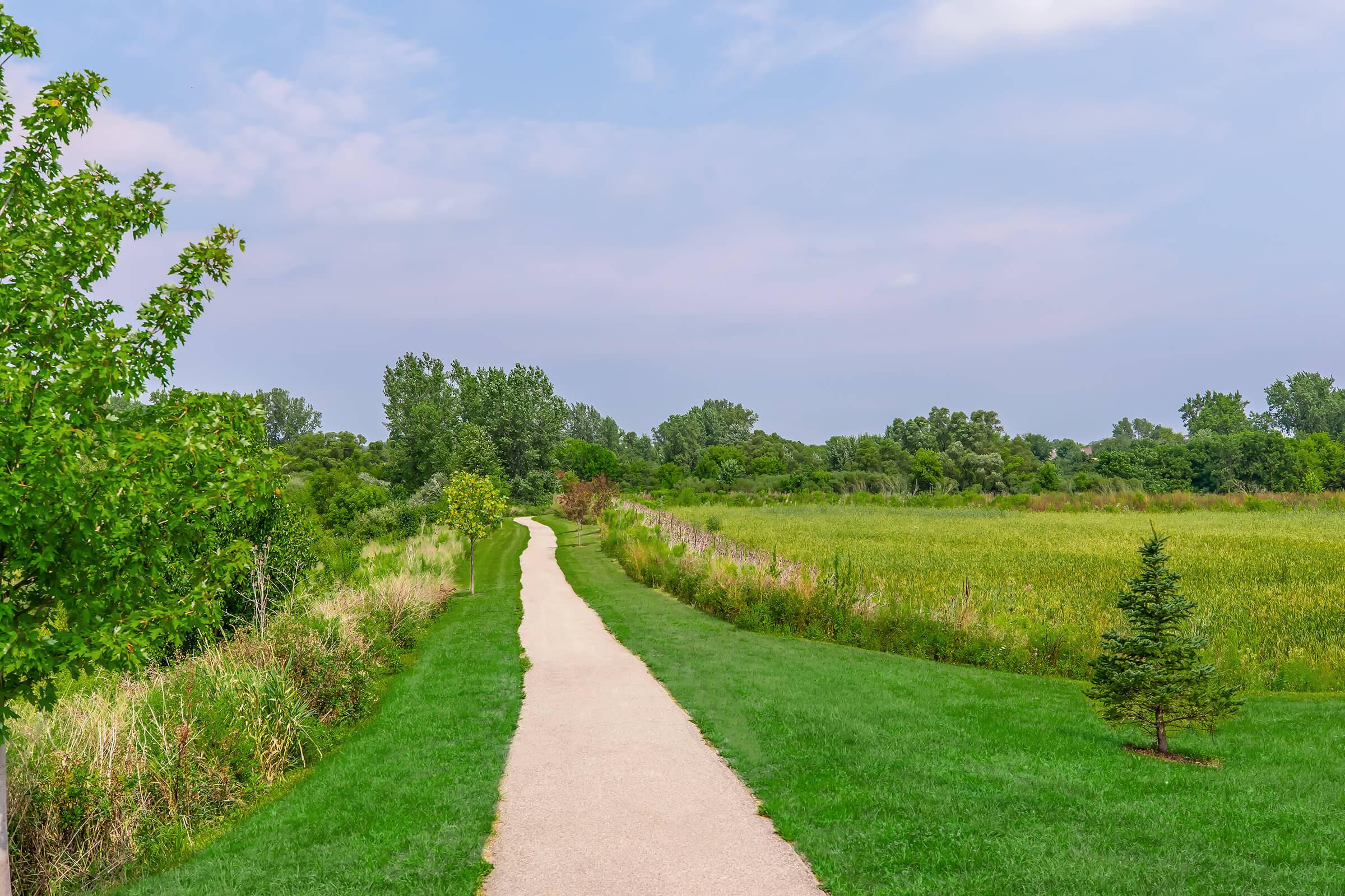 A winding path lined with greenery leads through a serene landscape of lush grass and trees, with a field of tall grass on one side. The sky above is partly cloudy, creating a peaceful and inviting outdoor scene.