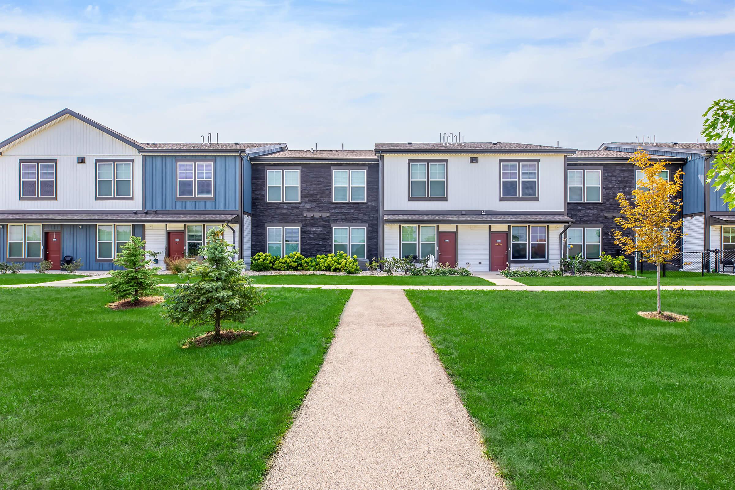A row of modern townhouses with a landscaped front yard featuring small trees and shrubs. A paved walkway leads through the green grass to the entrance of each unit. The buildings have a mix of blue and gray siding, with large windows and contrasting doors. The sky is clear and bright.