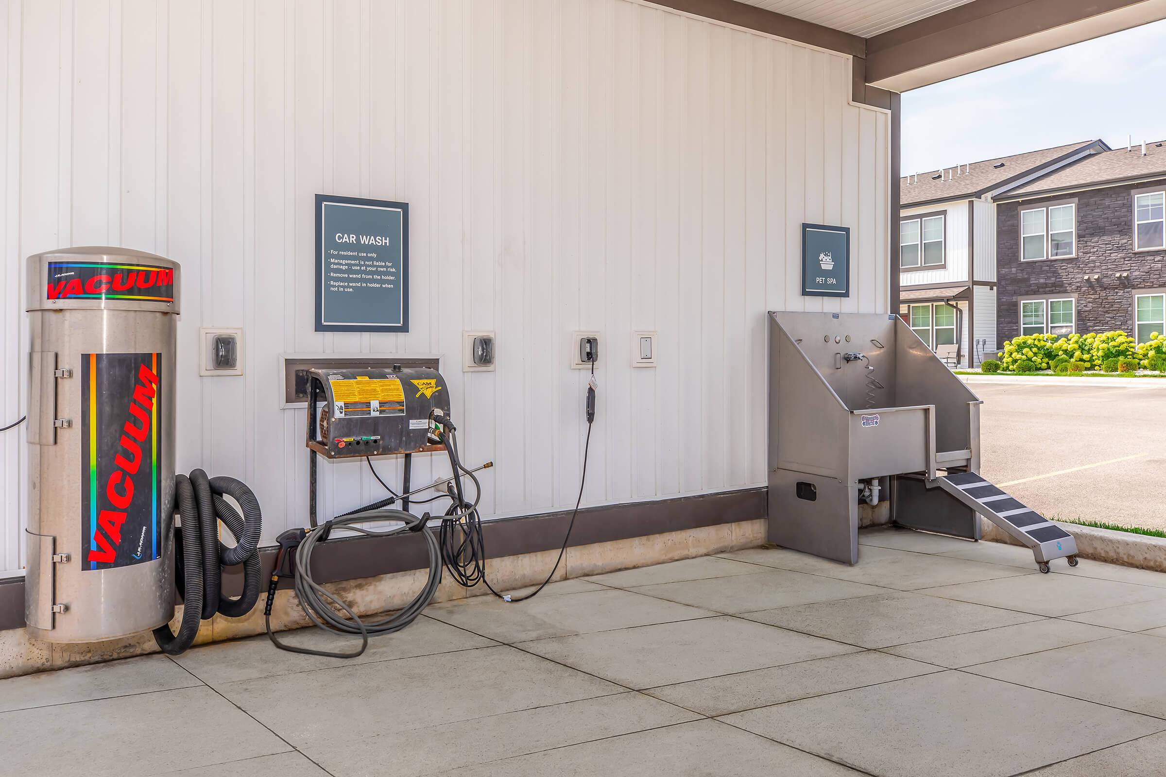 Self-service car wash area featuring a vacuum station and a wash bay. The vacuum has a bold "VACUUM" label, while the wash bay includes hoses and a sign indicating its use. The building has a clean, modern look with nearby residential units visible.