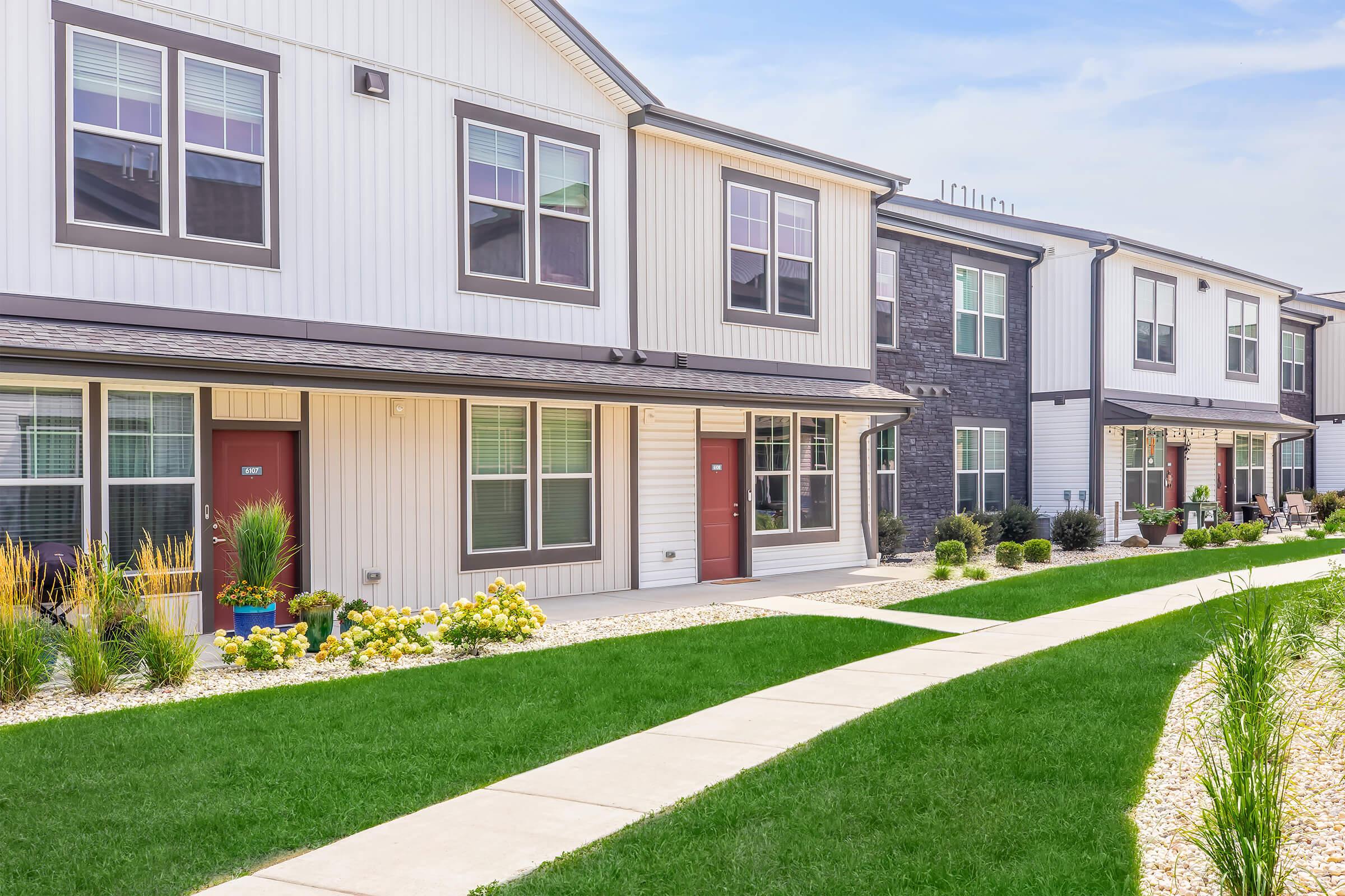 A row of modern townhouses with beige and dark gray siding, featuring red front doors and large windows. Well-maintained landscaping includes a green lawn with small shrubs and flowers, and a concrete walkway leading to the entrances of the homes. Bright blue sky in the background.